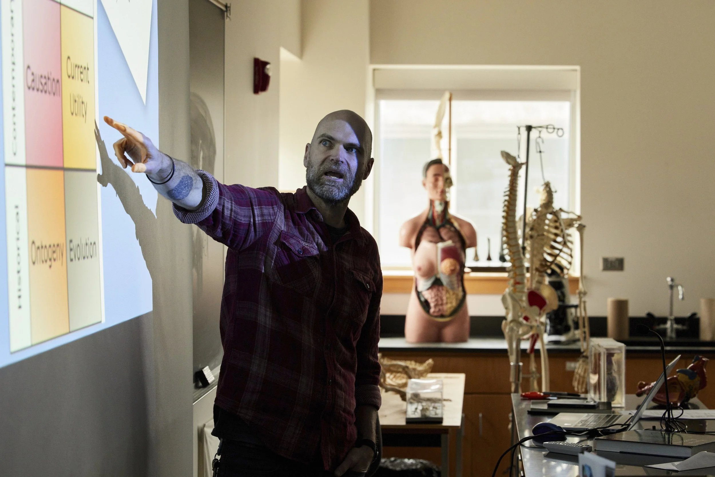 A man with a beard and a tattoo on his arm pointing at a presentation slide in a classroom, with a medical mannequin and skeleton in the background.