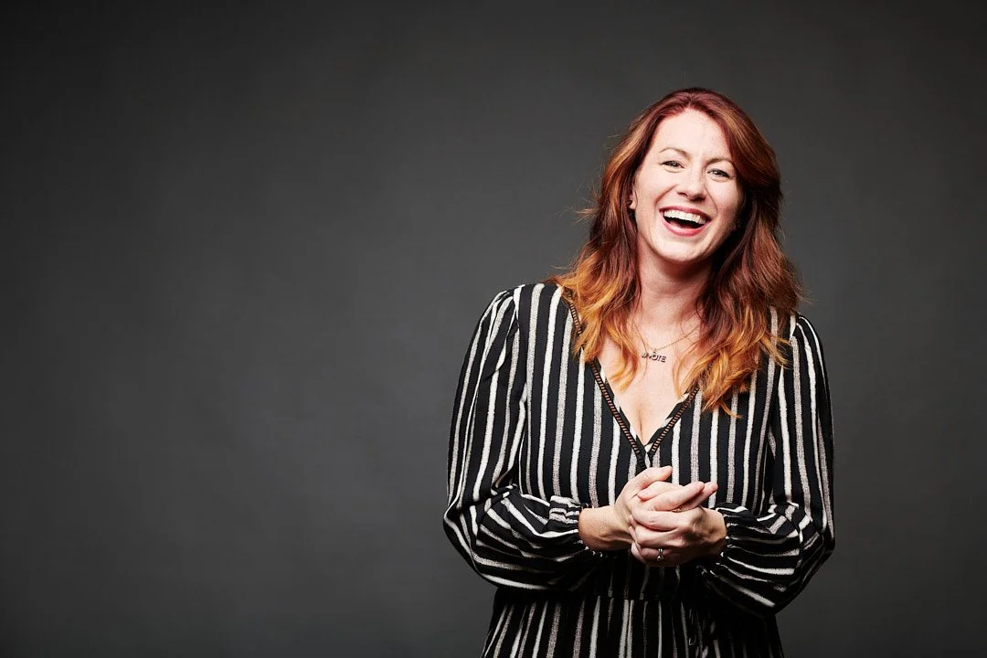 A woman with red hair and a black-and-white striped shirt laughing with her hands clasped in front of her.