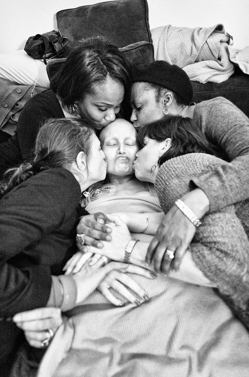 A woman with a bald head is lying on a bed and surrounded by four of her friends who are kissing her face and head and embracing her.  