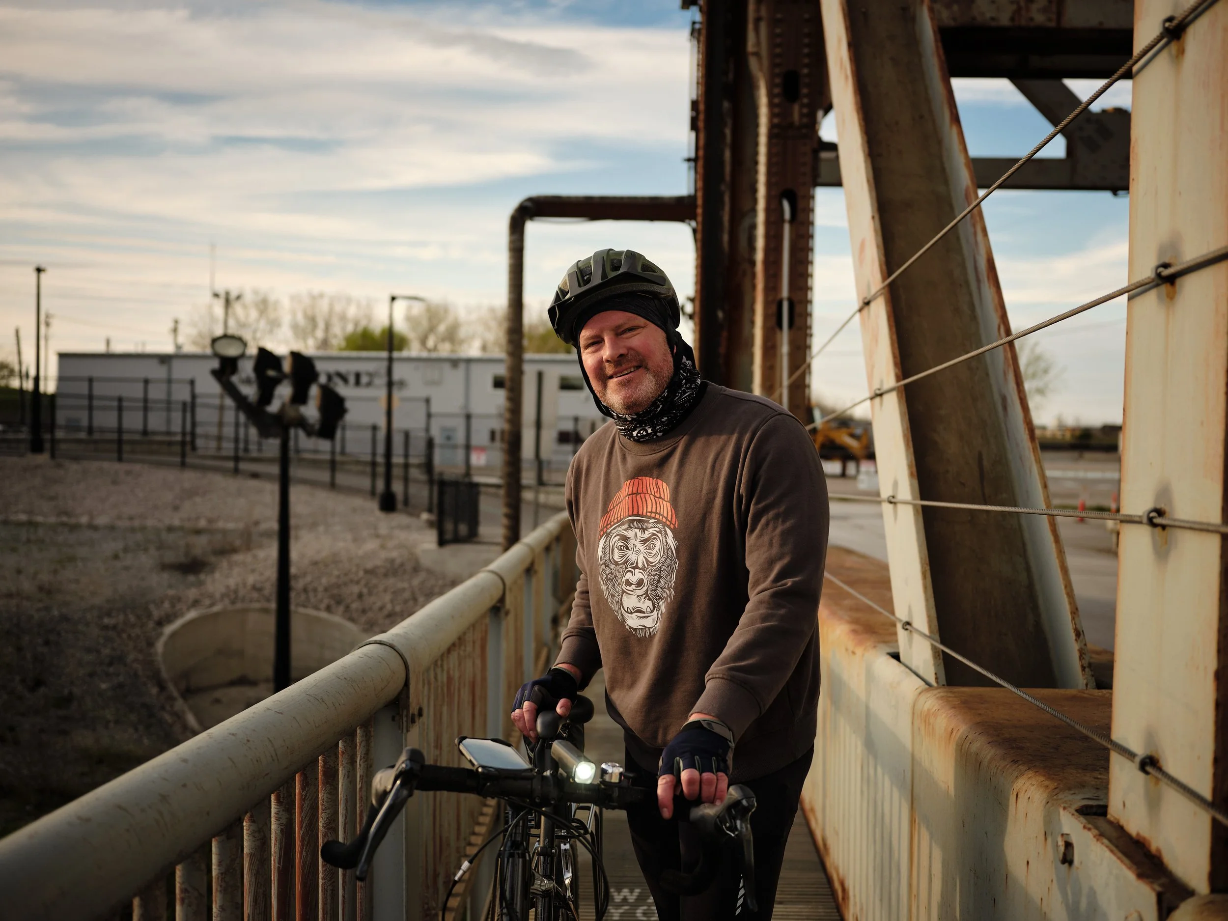 A man wearing a black helmet and brown sweatshirt with a gorilla face graphic, standing next to a bicycle, on a rusted metal bridge outdoors during twilight.