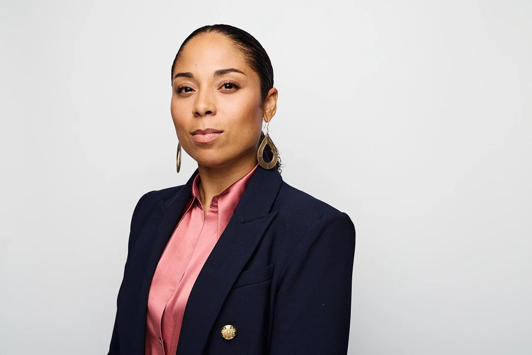 A woman with dark hair pulled back, wearing a dark blazer with gold buttons, a pink satin blouse, and gold earrings, standing against a plain gray background.