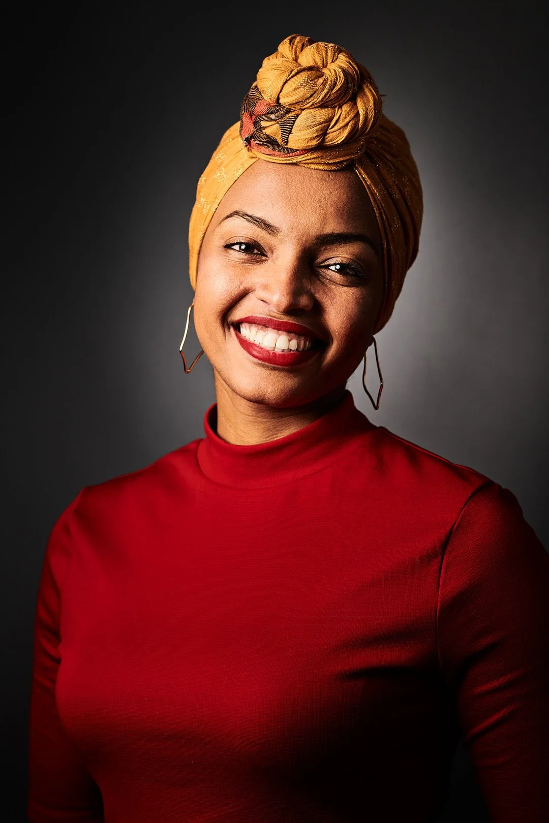 Smiling woman wearing a yellow headwrap and red top against dark background.
