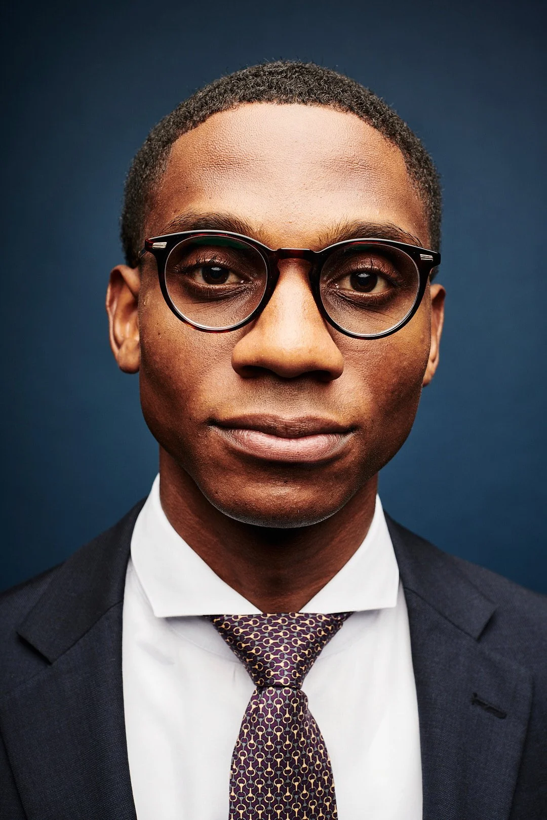 Close-up portrait of a young African American man wearing glasses, a dark suit, white shirt, and a patterned tie against a solid dark background.