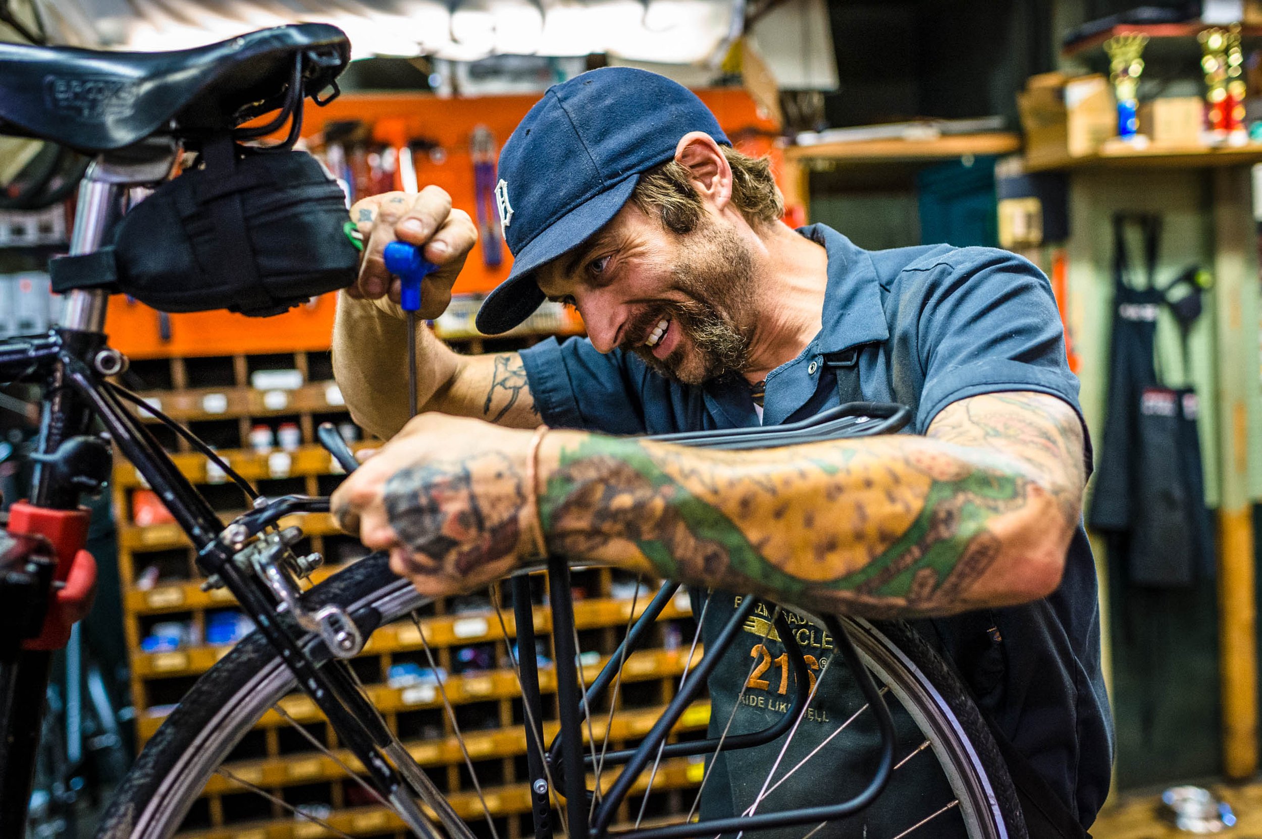 A man with tattoos, wearing a blue cap and shirt, working on repairing a bicycle in a workshop, smiling as he uses tools.