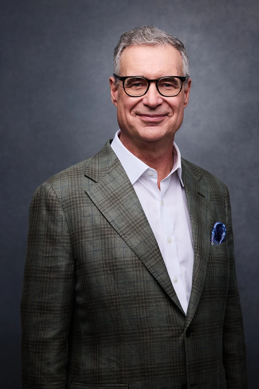 A middle-aged man with gray hair and glasses, dressed in a plaid blazer, white shirt, and pocket square, standing against a dark background and smiling.