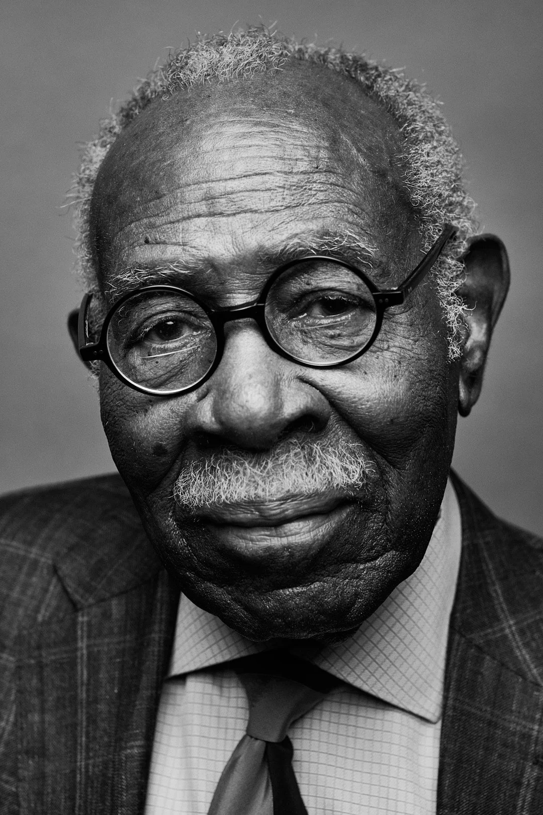 A black-and-white portrait of an elderly man with glasses, gray mustache, and hair, wearing a checkered jacket, shirt, and tie.