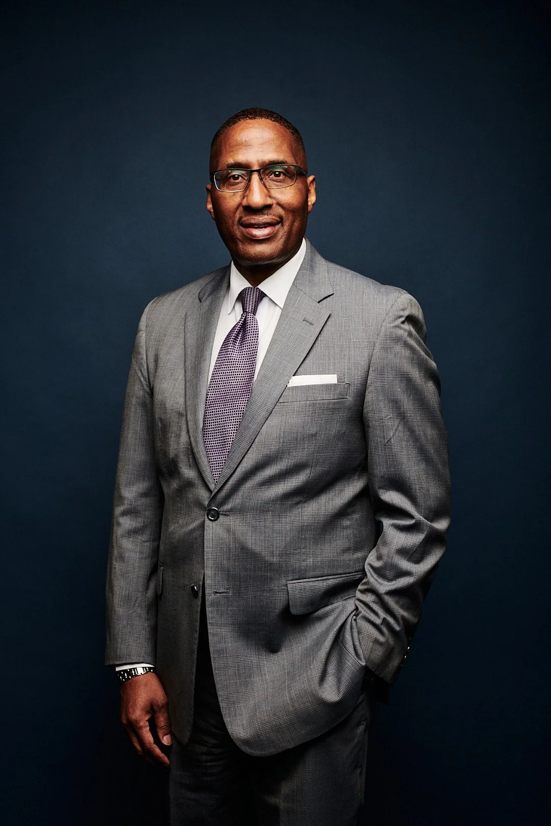 A man in a gray checkered suit, white shirt, and purple tie standing against a dark blue background, wearing glasses and a watch.