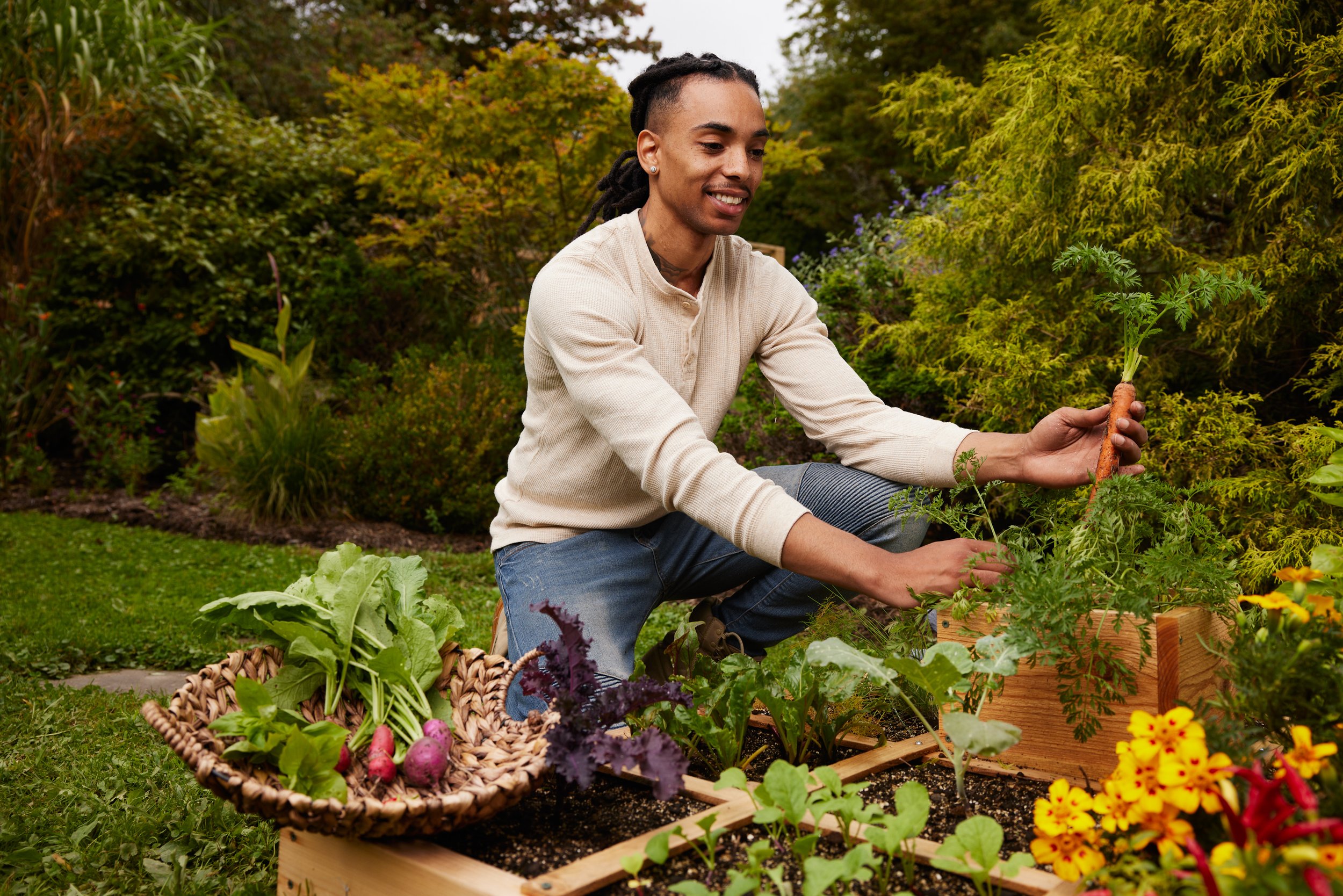 Person gardening in a backyard with a variety of vegetables and flowers, holding a carrot.