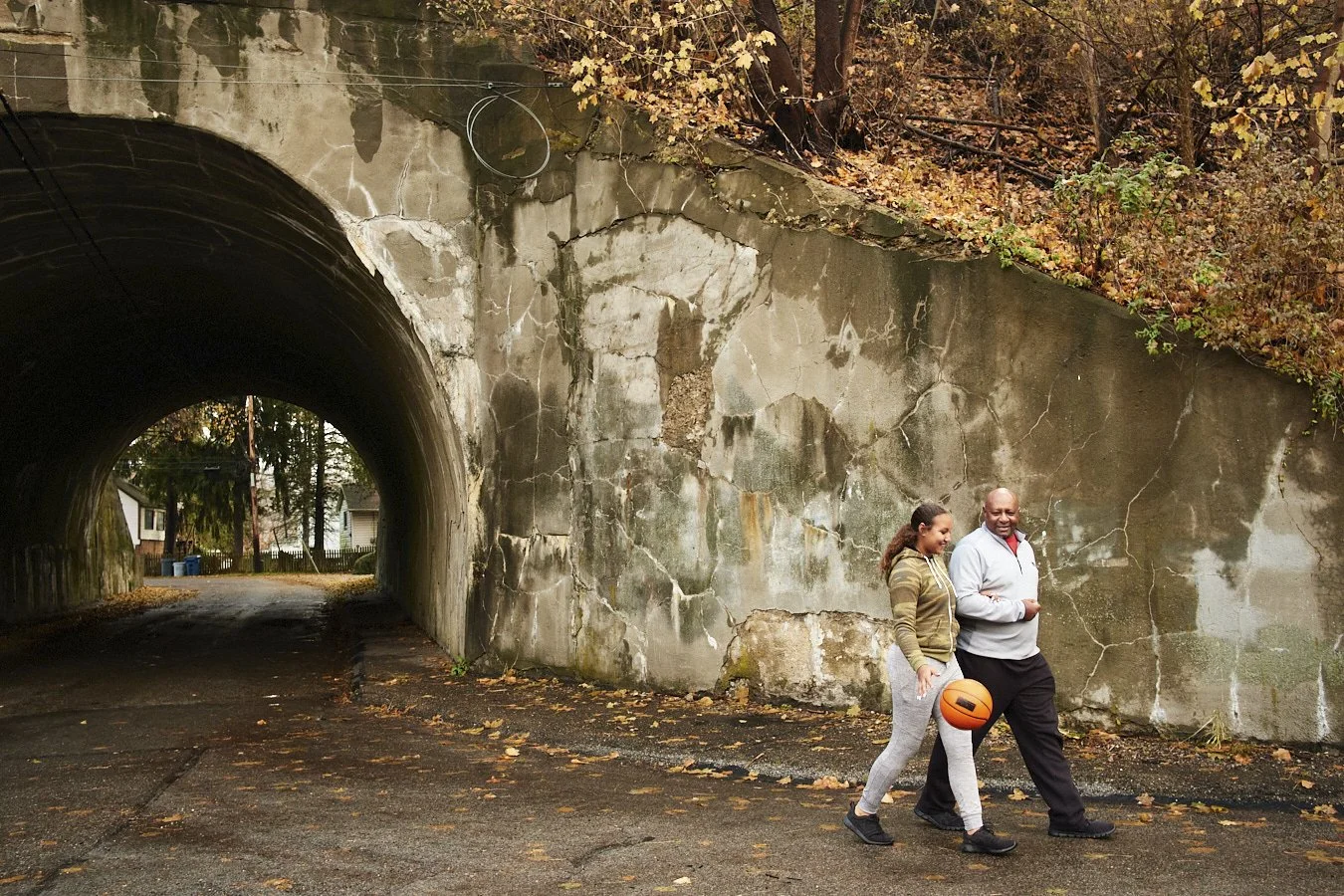 A man and woman walk on a sidewalk under a concrete tunnel with autumn leaves strewn on the ground and trees with fall foliage in the background. The man is holding an orange basketball.