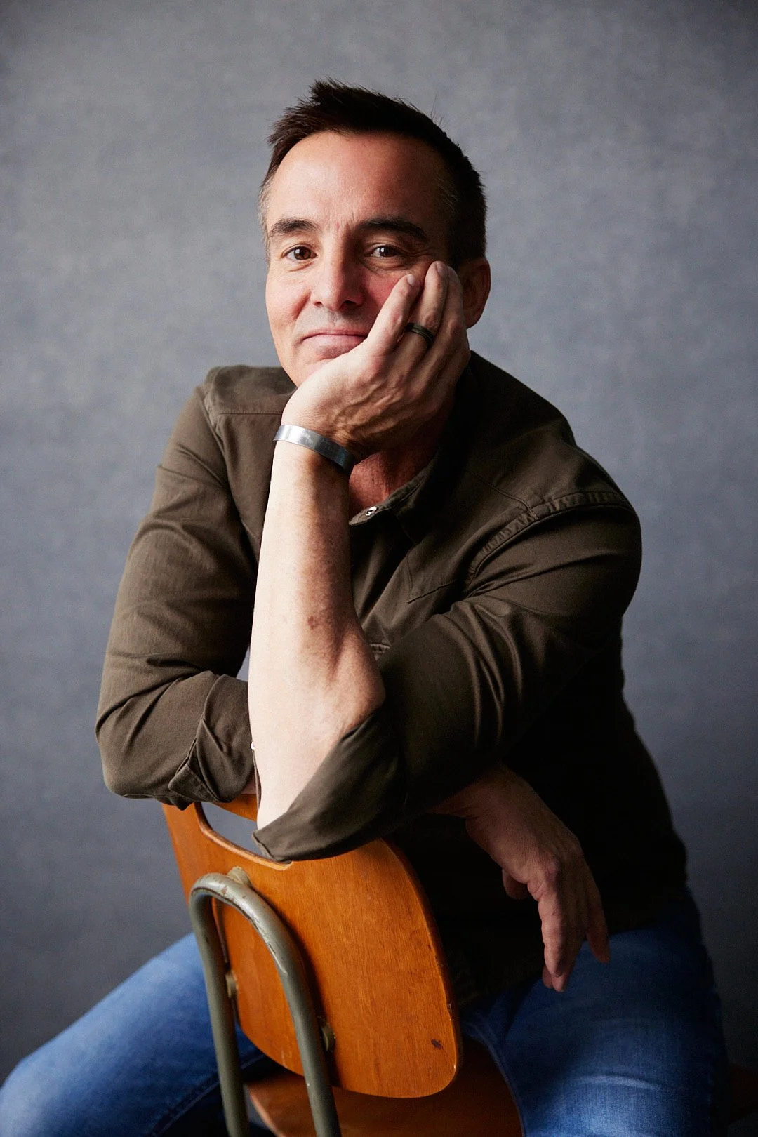 A man with short dark hair, wearing a brown shirt, sitting on a wooden chair with a metal backrest, resting his head on his hand, looking at the camera with a thoughtful expression.