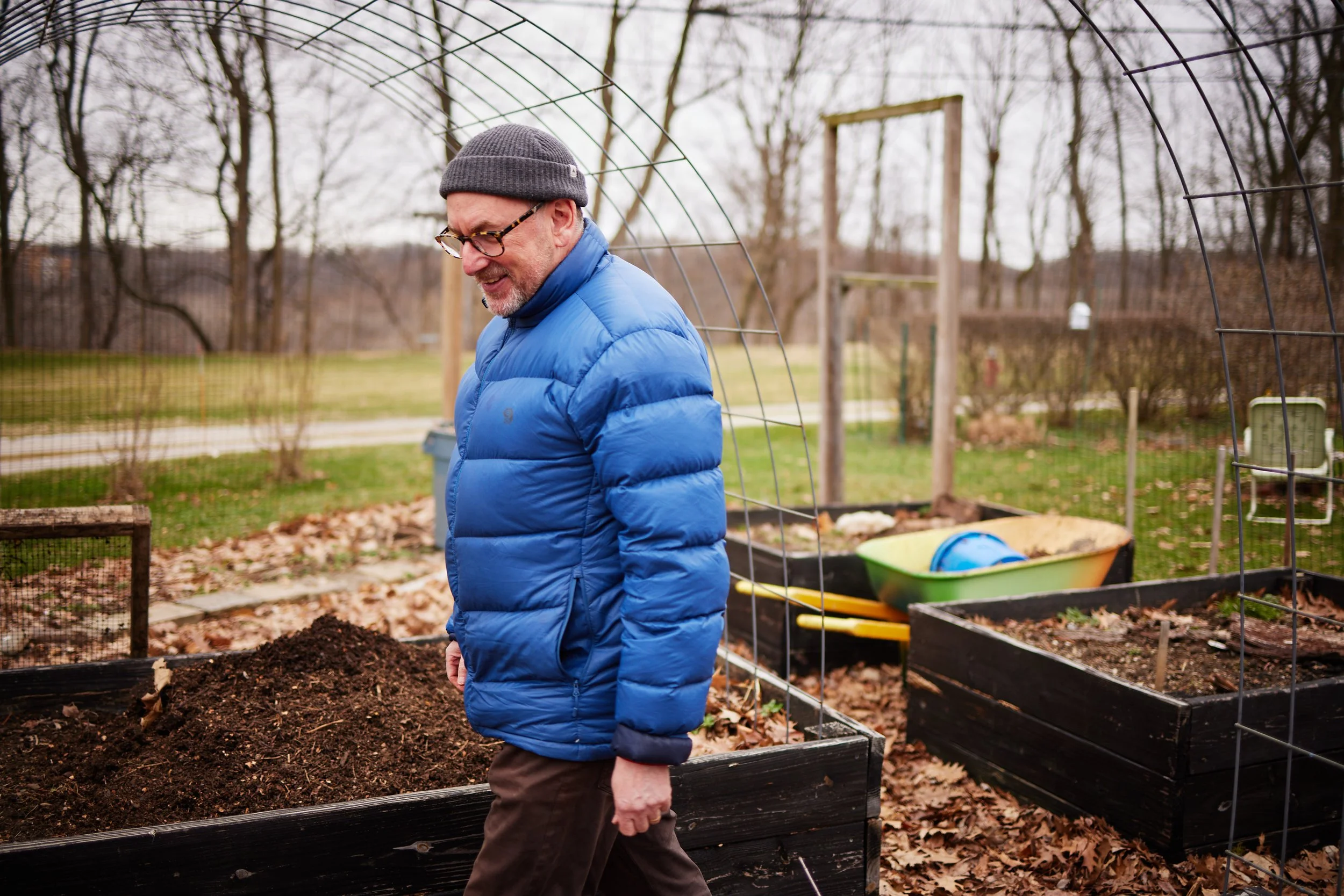 A man in a gray beanie, glasses, and a blue puffer jacket walking past a garden bed with dark soil in an outdoor garden or backyard during fall or winter.