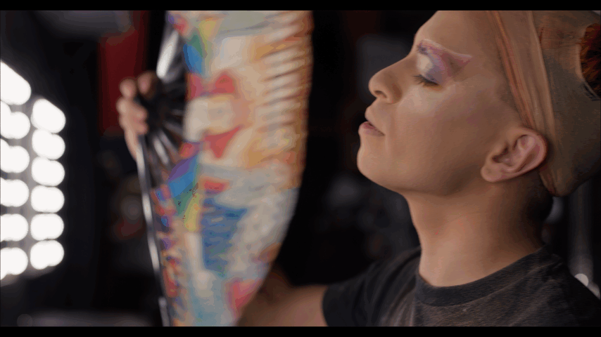 Child with face makeup and hair wrapped in a cloth, holding a colorful umbrella, with eyes closed, in a makeup or dressing room.