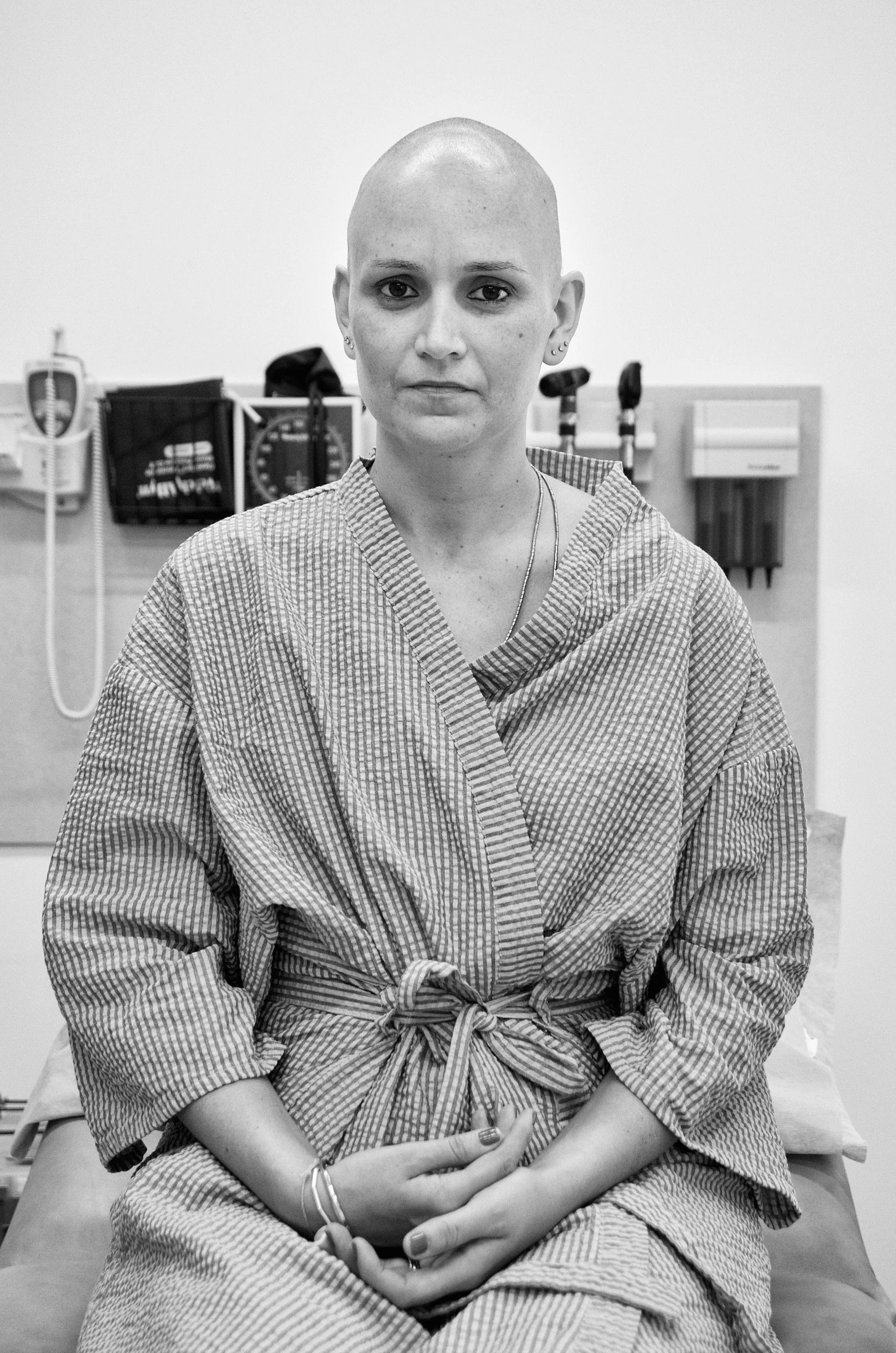 A woman with a shaved head sitting on a hospital bed, wearing a hospital gown, in a medical examination room.