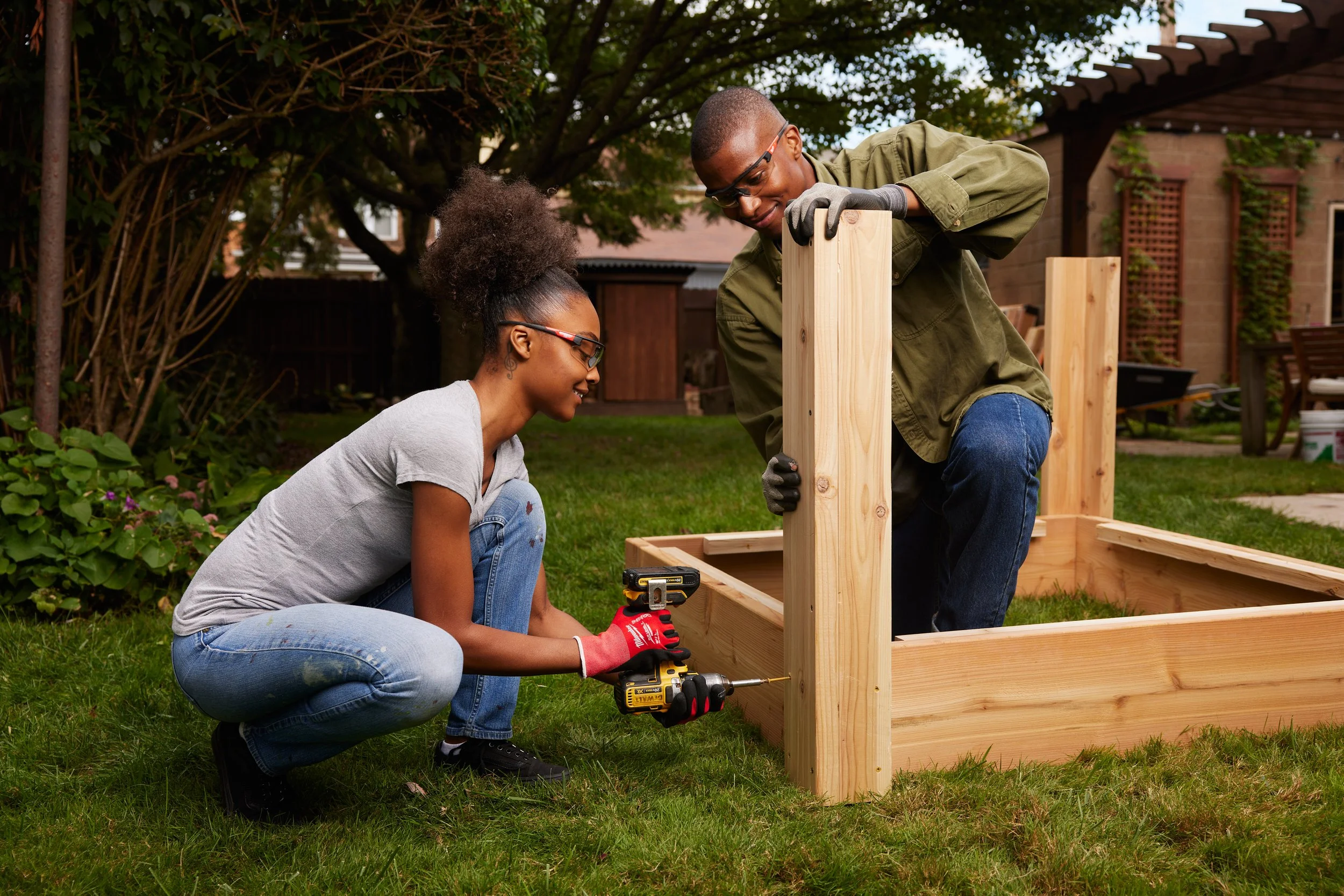 Two people building a wooden structure outdoors, one is kneeling with a drill, the other is holding a wooden post. They are working on a gardening or outdoor project in a backyard.
