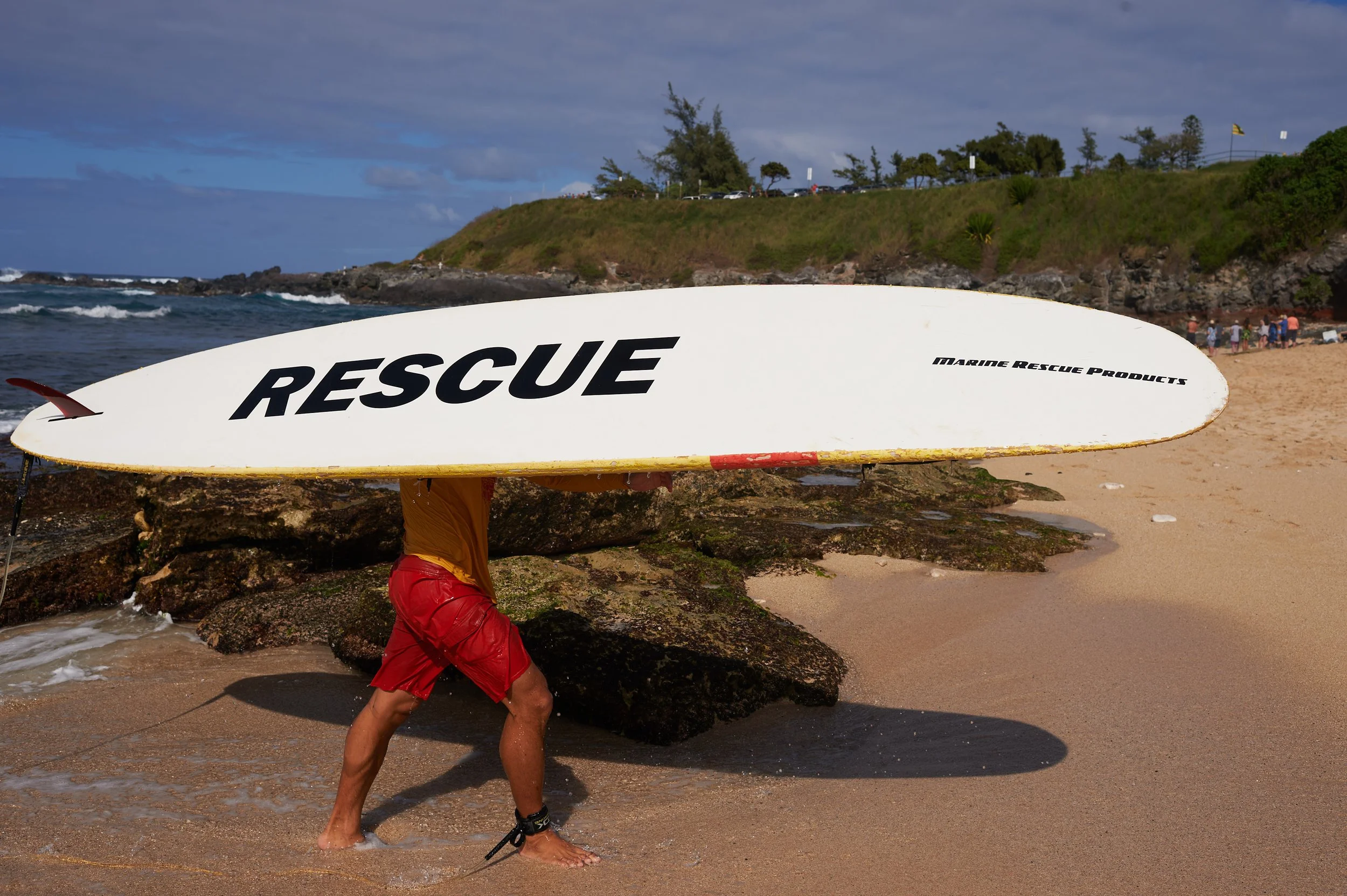 Person in red shorts carrying a white rescue surfboard labeled "RESCUE" on a sandy beach with rocks, ocean waves, and a grassy hill in the background.