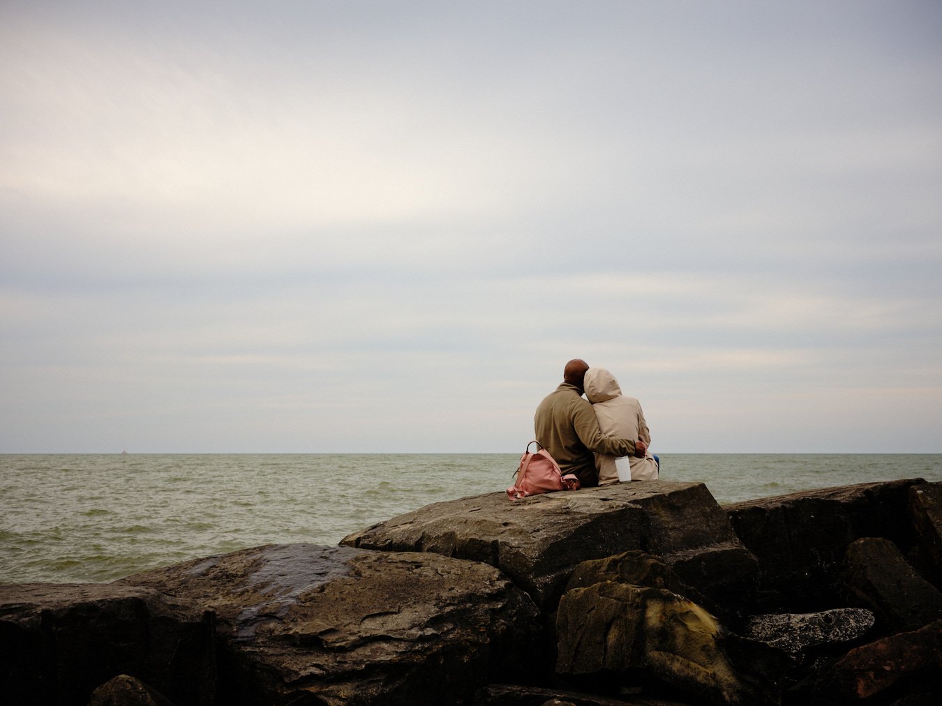 A couple sitting on rocks by the ocean, embracing and looking at the water, with bags beside them under a cloudy sky.