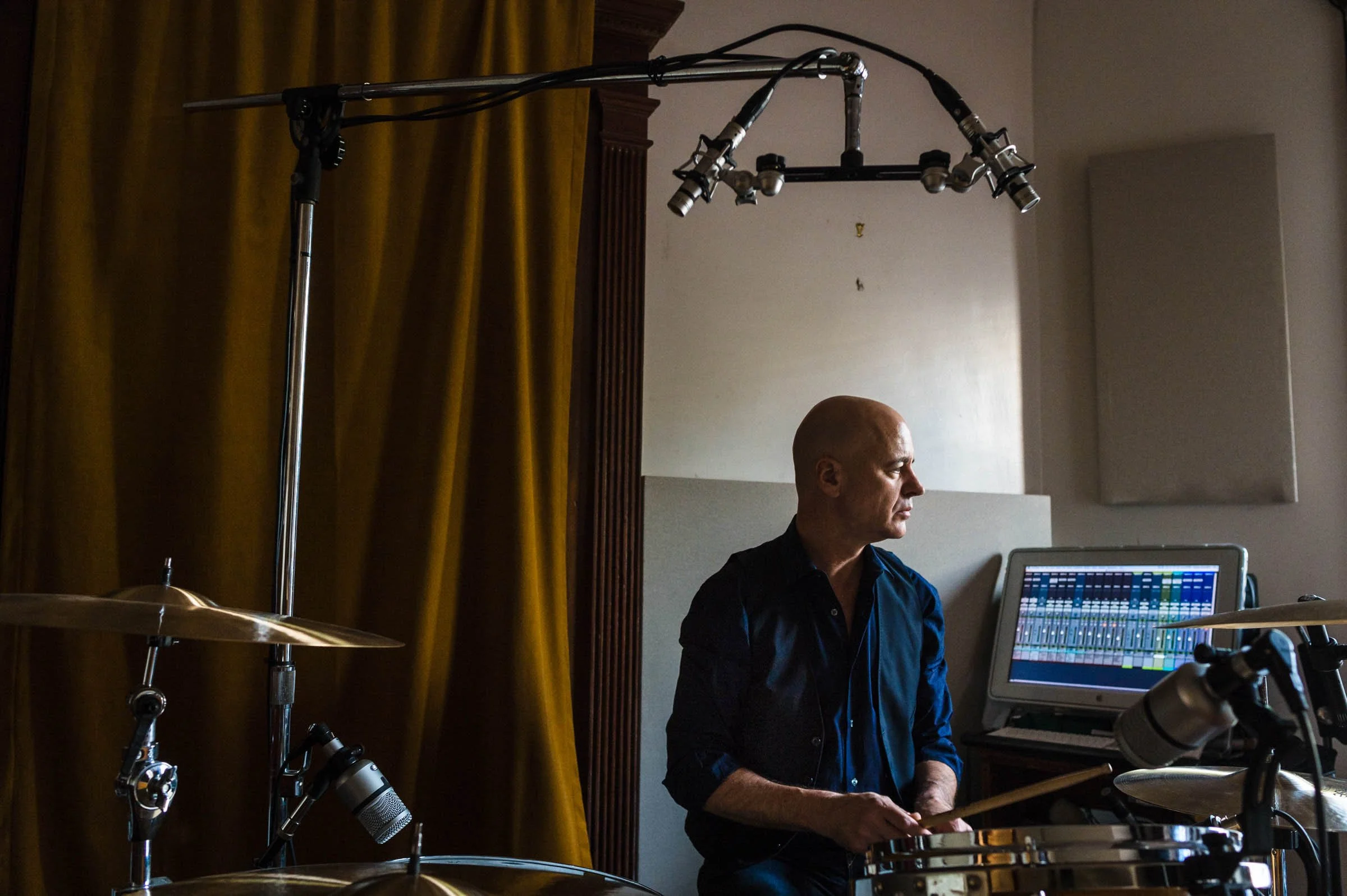A man with a bald head playing drums in a recording studio, with a computer monitor displaying audio mixing software in the background.