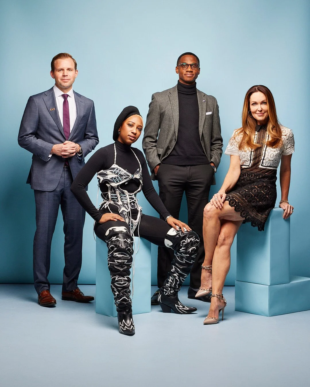Group of five diverse professionals posing against a plain blue background, with some seated on light blue blocks, dressed in business and fashion-forward clothing.