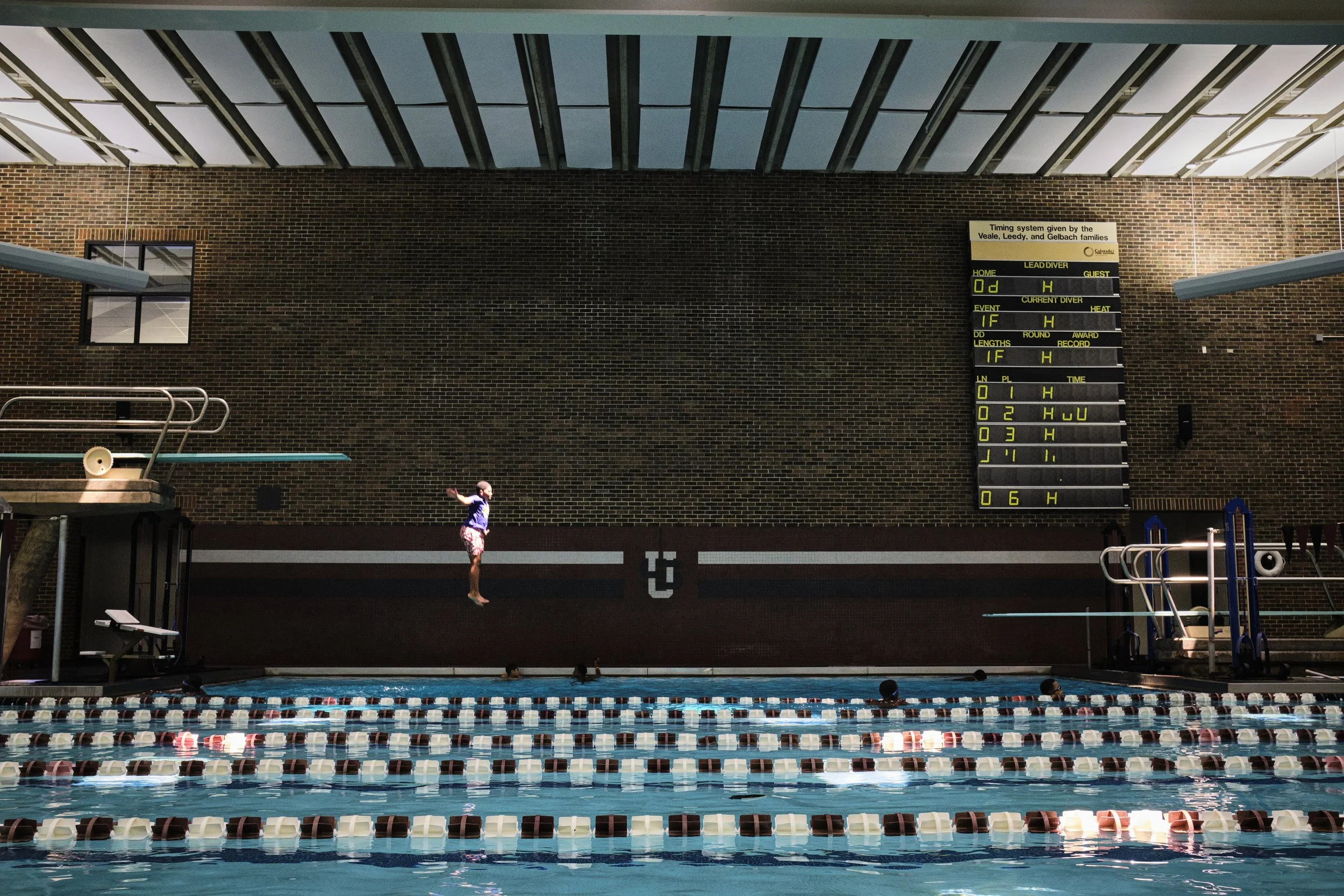 An indoor swimming pool with a young girl jumping into the water, and a scoreboard on a brick wall showing race data.