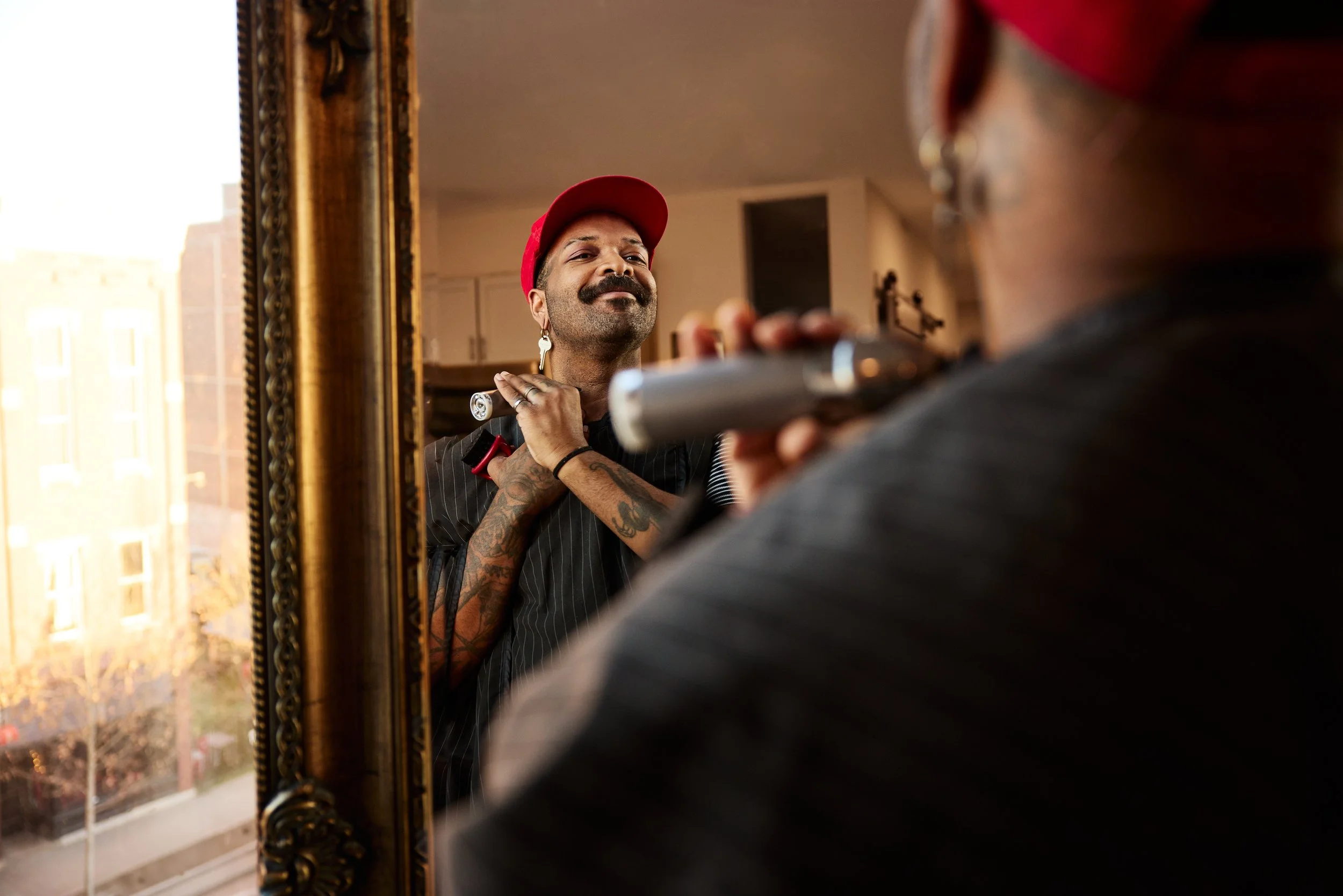 A person with tattoos and earrings is blow-drying their hair while looking in a mirror, wearing a red cap and dark striped clothing, in a well-lit room with a window showing city buildings outside.