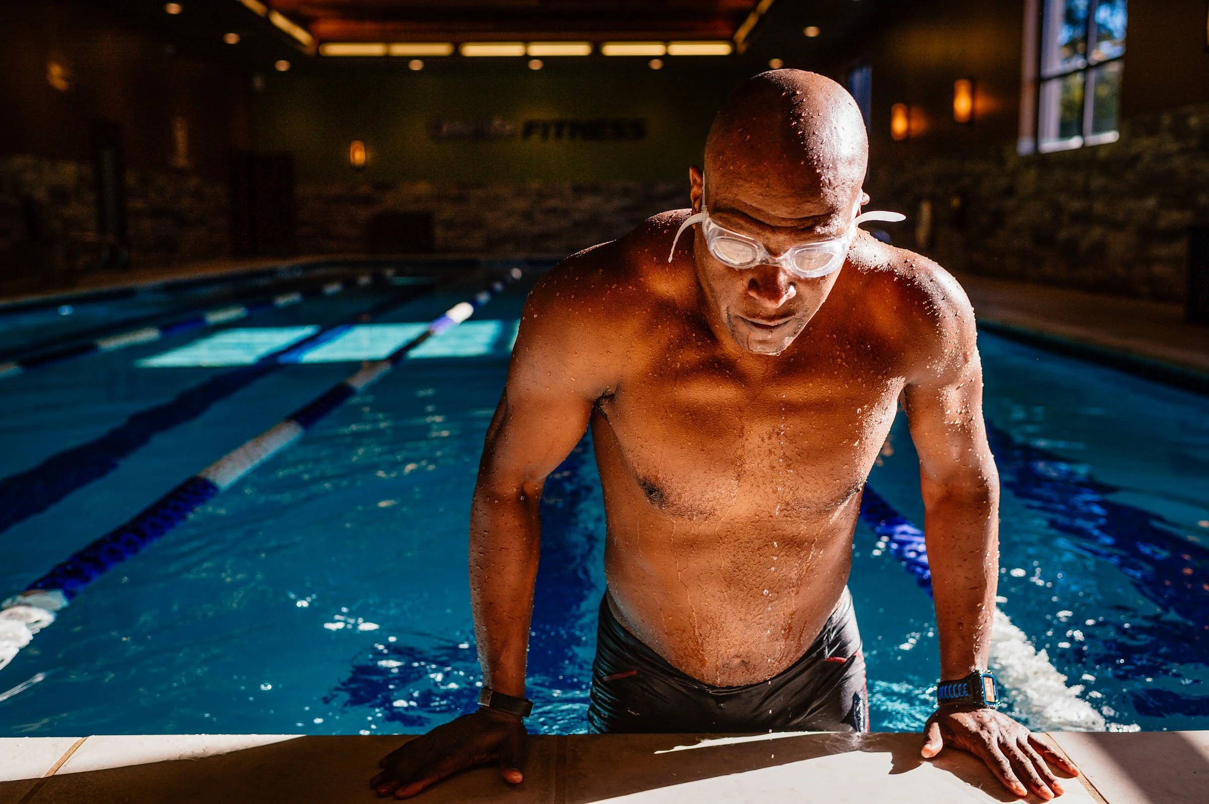 A man emerges from a swimming pool, wearing goggles and a smartwatch, with a serious expression, in an indoor pool setting.