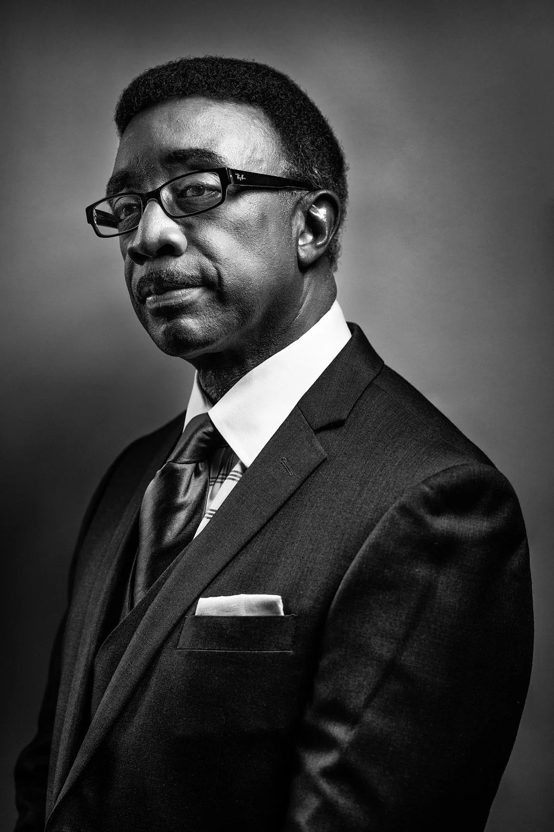Black and white portrait of a man wearing glasses, a suit, tie, and pocket square, looking at the camera with a serious expression.
