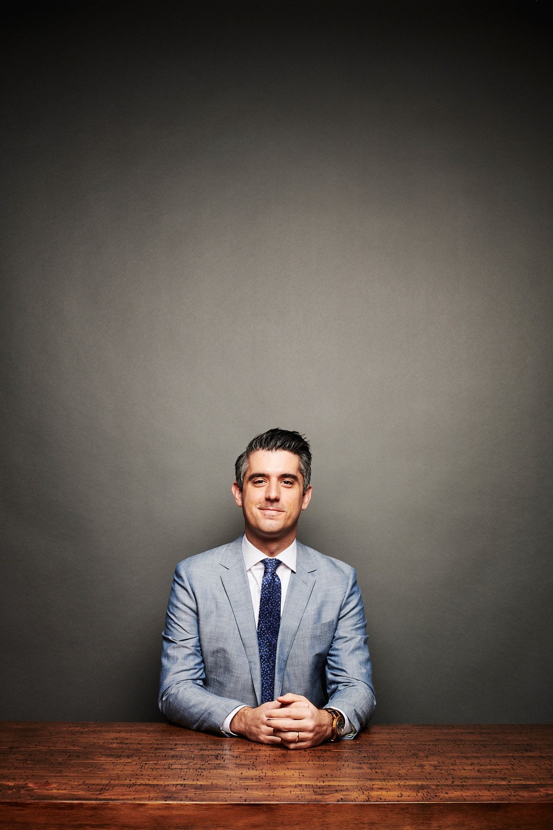 A man in a light gray suit and tie sitting at a wooden table against a plain gray background.