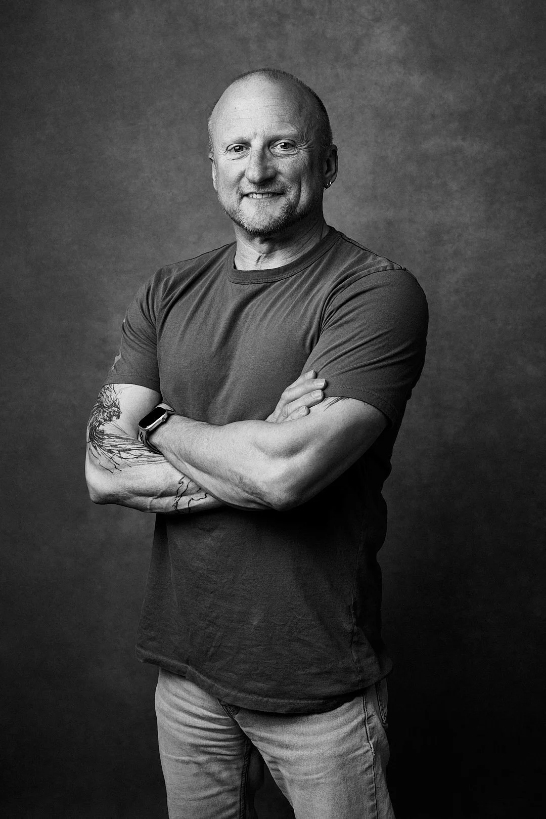 Black and white portrait of a smiling man with a beard, wearing a T-shirt and jeans, crossing his arms with a tattoo on his left arm.