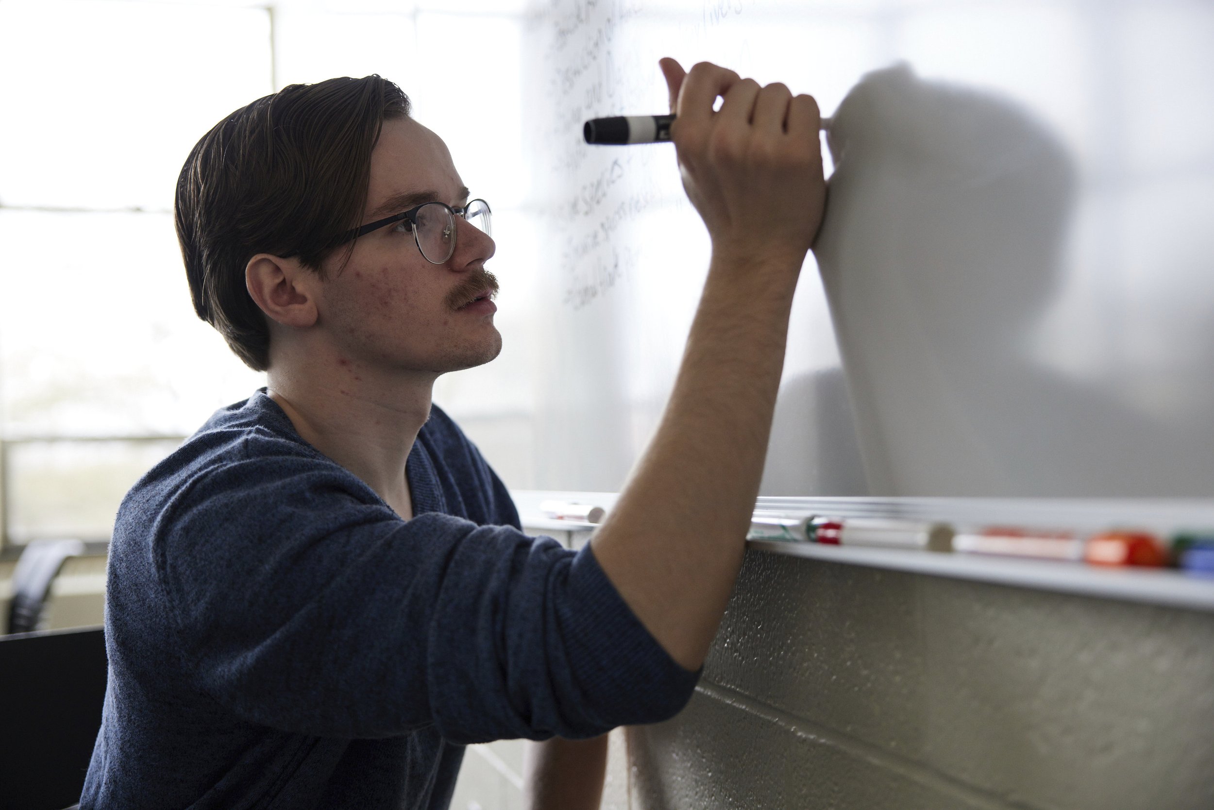 Young man with glasses writing on a whiteboard in a classroom.