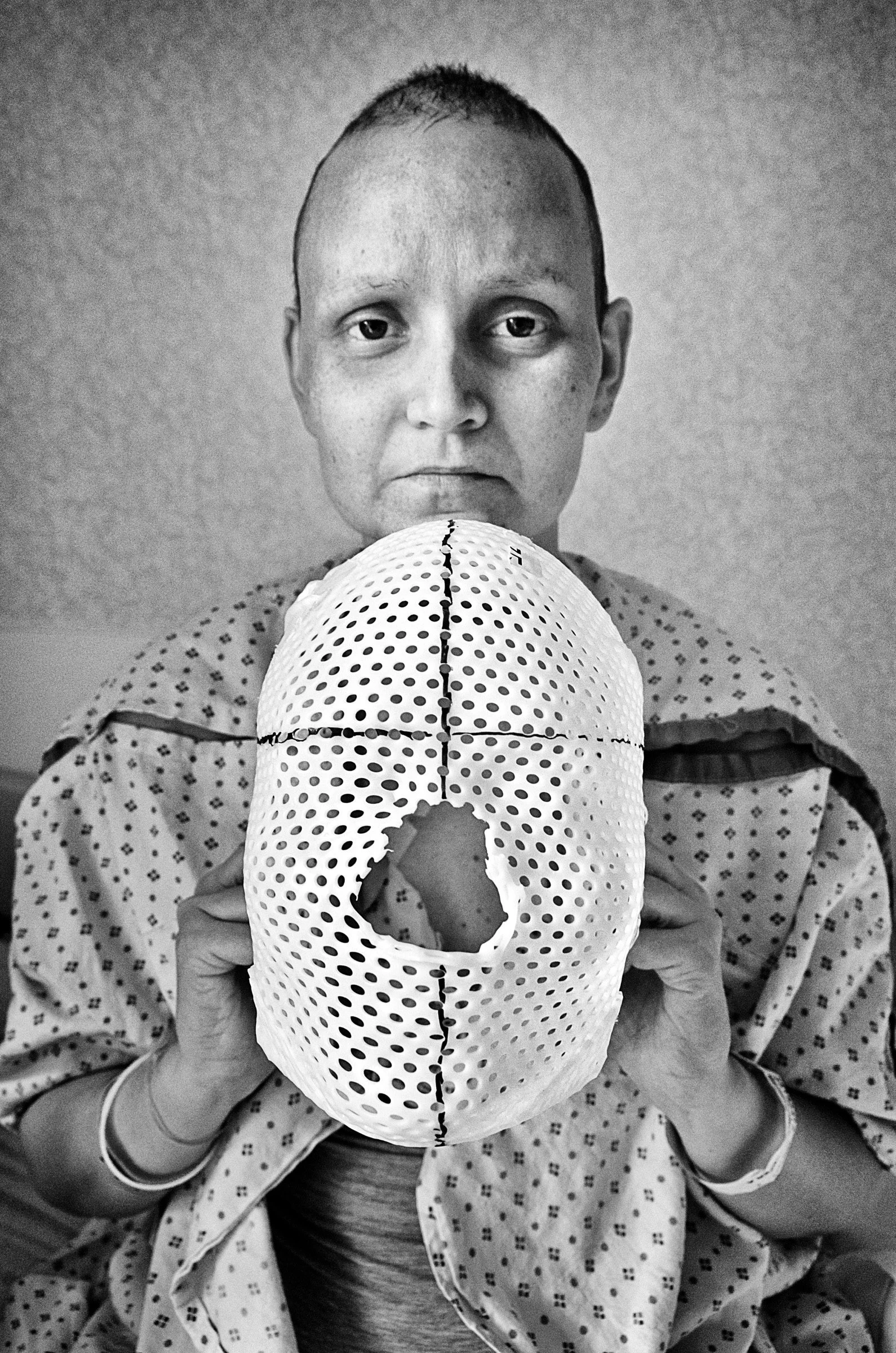 A person in a hospital gown holding a perforated mask resembling a skull in front of their face, with a serious expression.