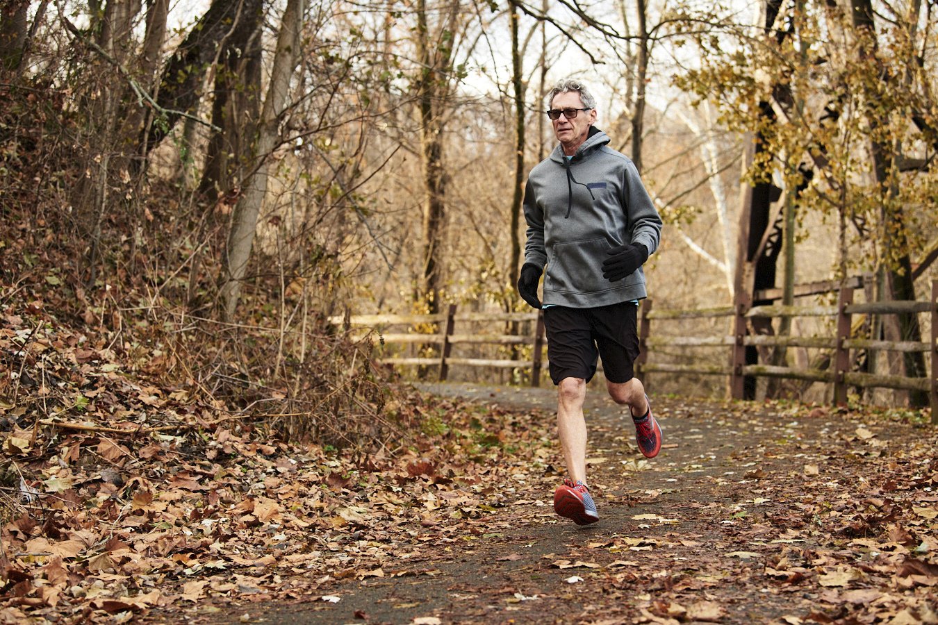 A man jogging alone on a leaf-covered trail in a forest with autumn trees in the background.