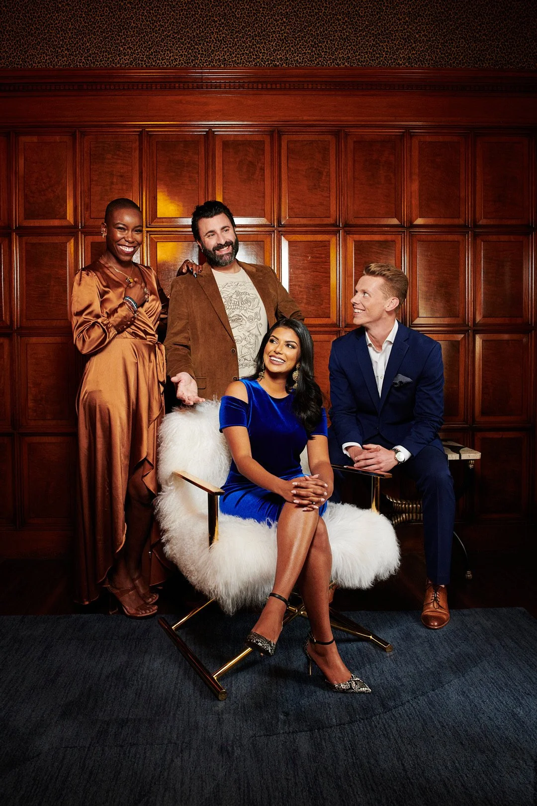 Four well-dressed people in a formal setting pose for a photo against a wooden-paneled wall. One woman is seated in a white fur-lined chair wearing a blue velvet dress with gold earrings, and three others stand around her, smiling and engaging with e