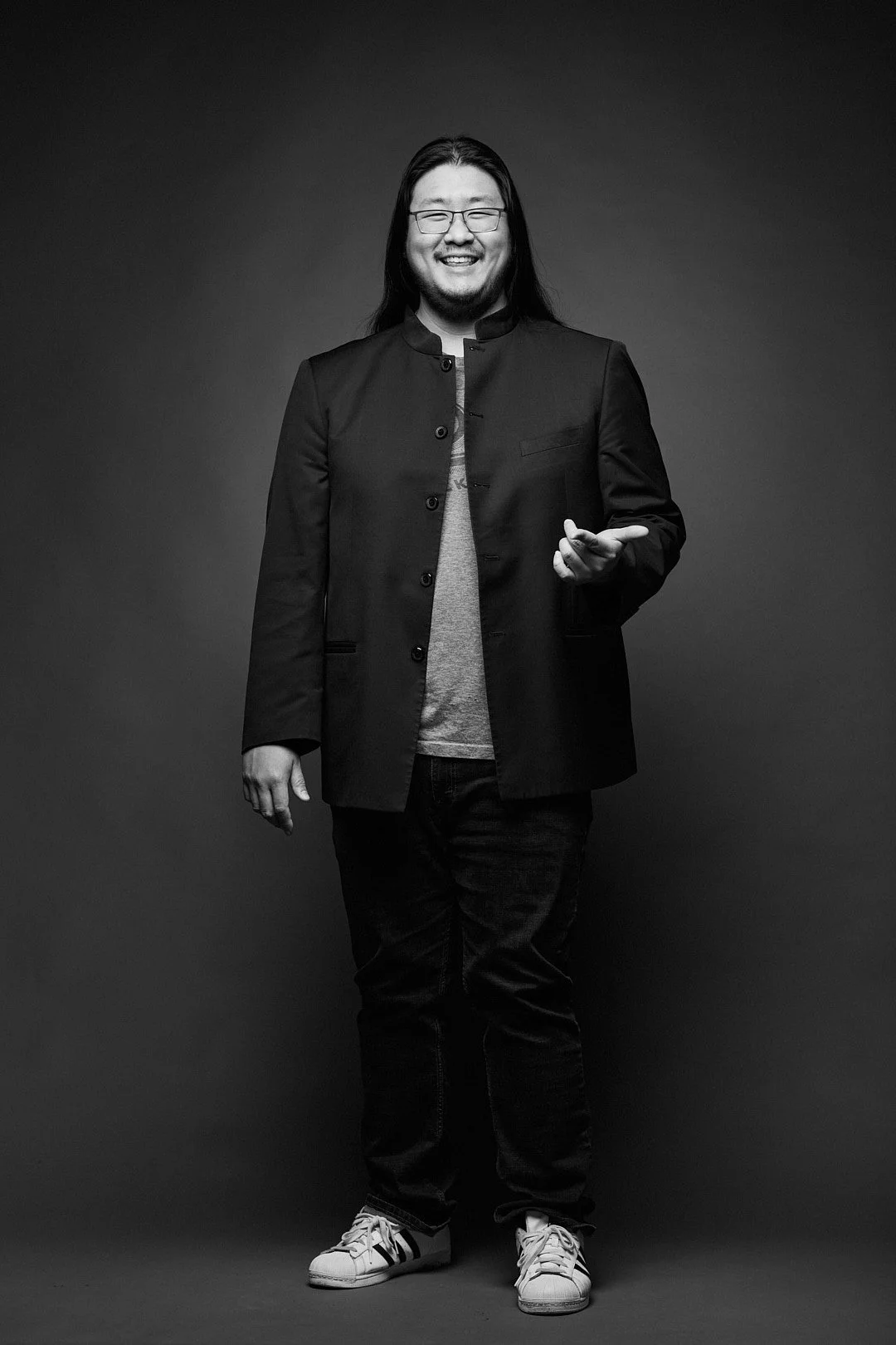 A smiling man with long hair, glasses, wearing a dark jacket, t-shirt, jeans, and sneakers, standing against a plain dark background.