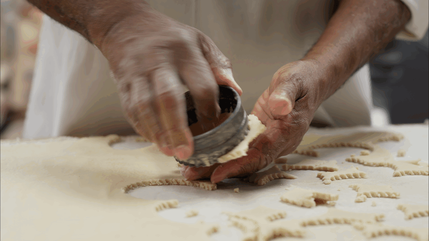 A person is using a round cutter to cut out shapes from a sheet of dough on a flat surface. The person's hands are covered in flour or dough. There are multiple dough shapes on the surface.