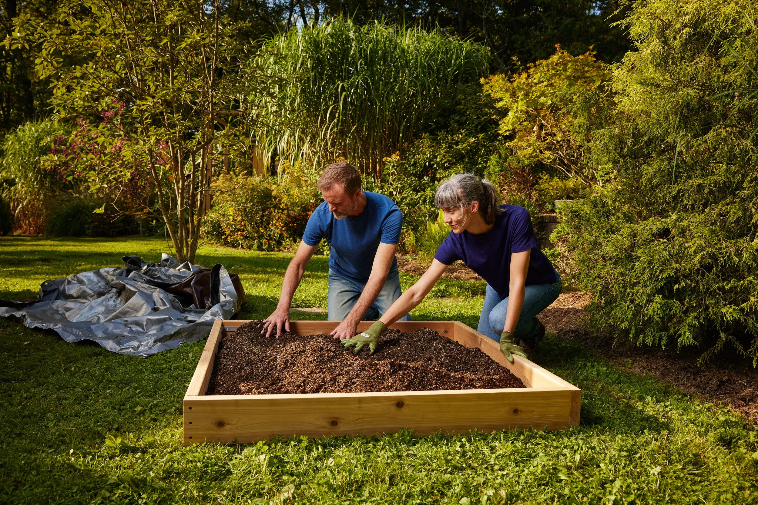 Two people working together in a garden, spreading soil or compost into a wooden garden bed, with trees and plants in the background.