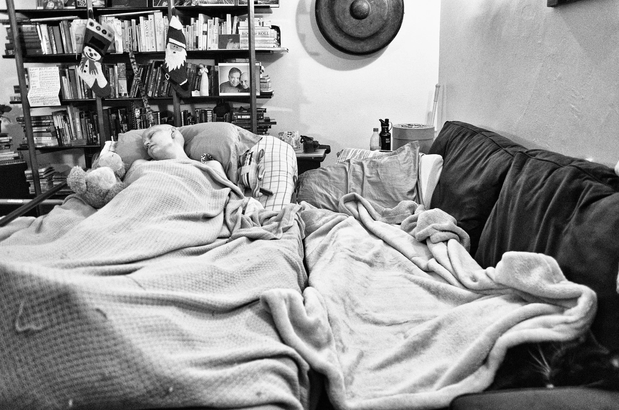 A person sleeping in bed with blankets, surrounded by books on a bookshelf, a teddy bear, and various personal items in a cozy room decorated with holiday stockings hanging from the bookshelf.