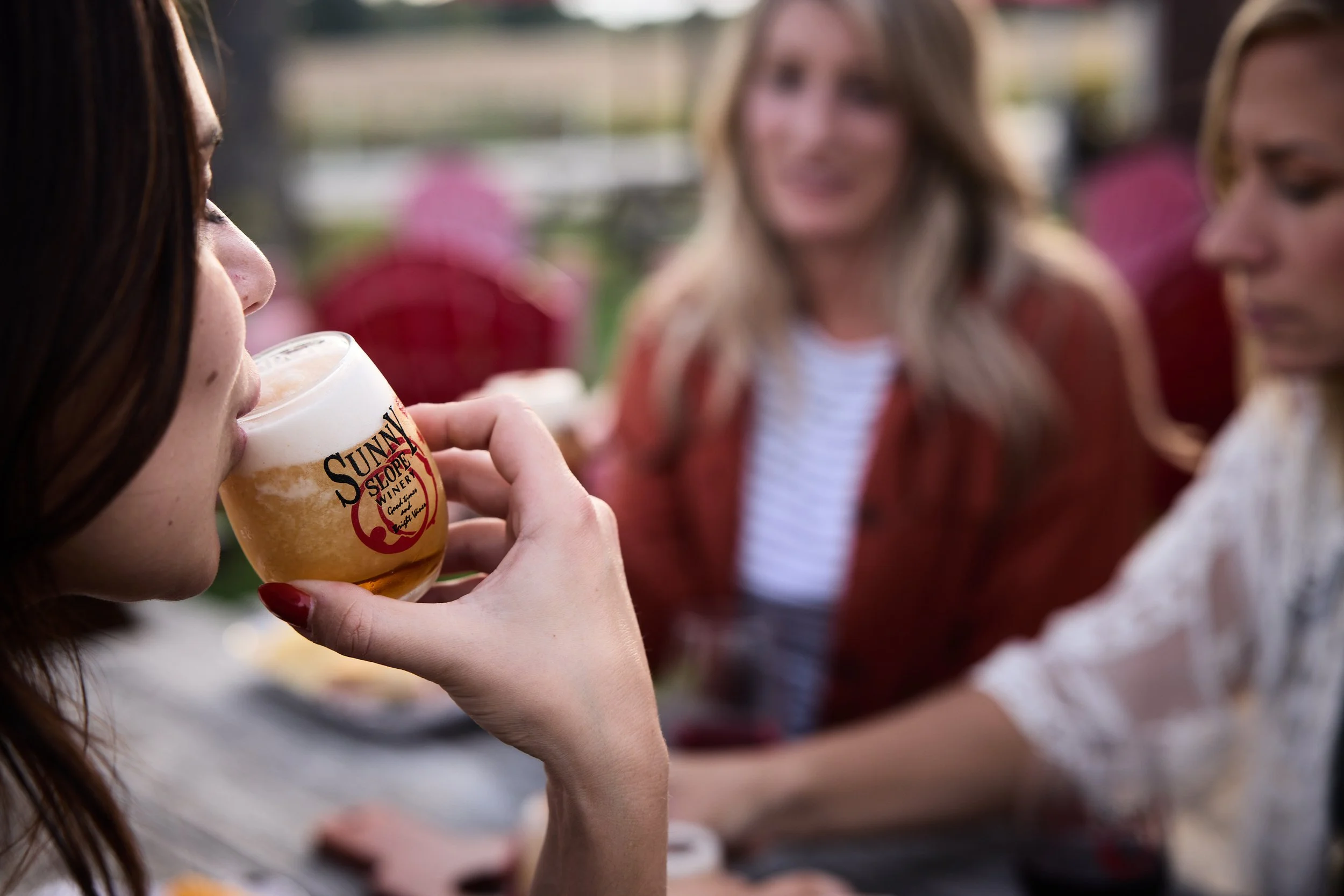 A woman with dark hair drinking beer at an outdoor gathering with other women in the background.