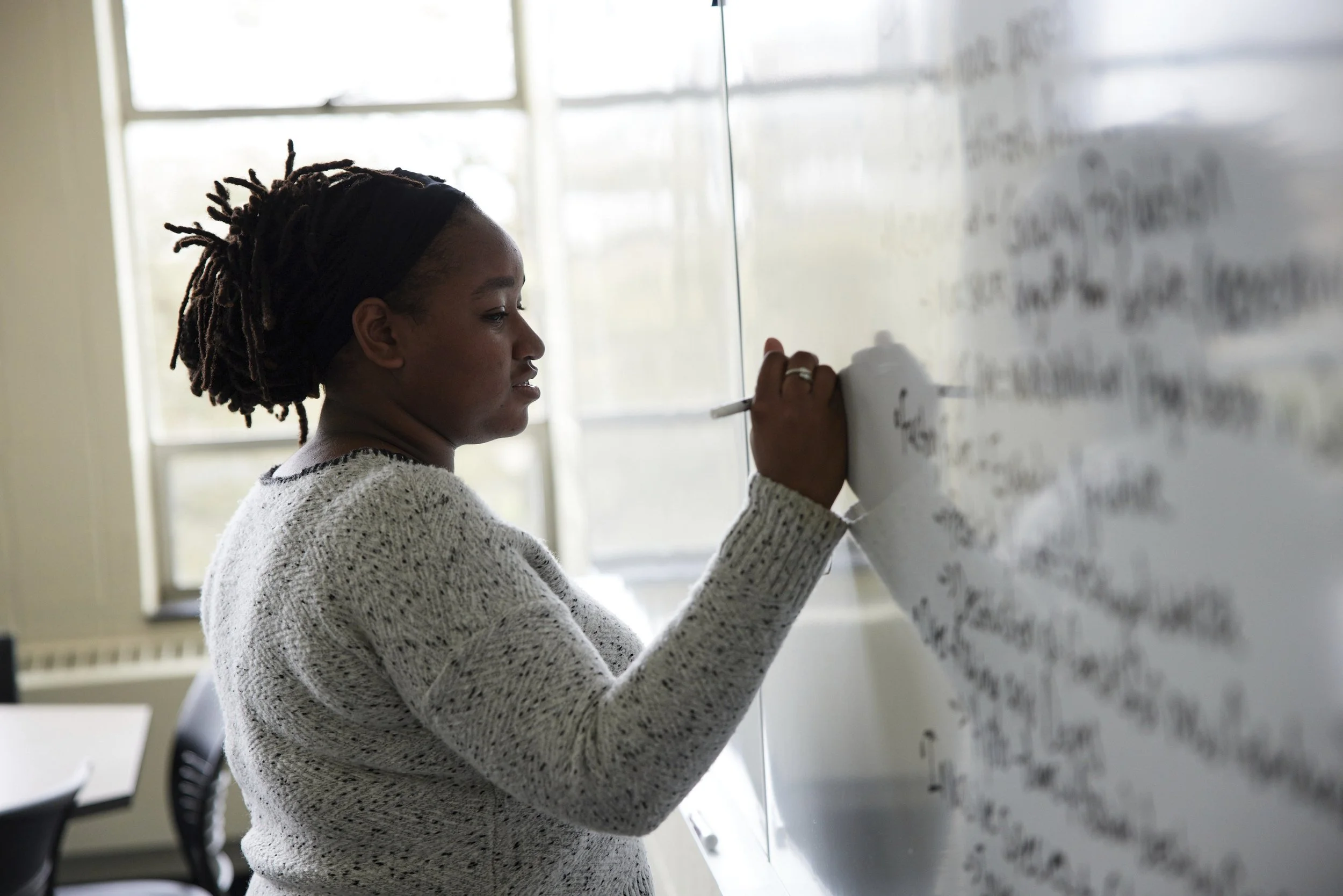 Woman writing on a whiteboard in a classroom with large windows.