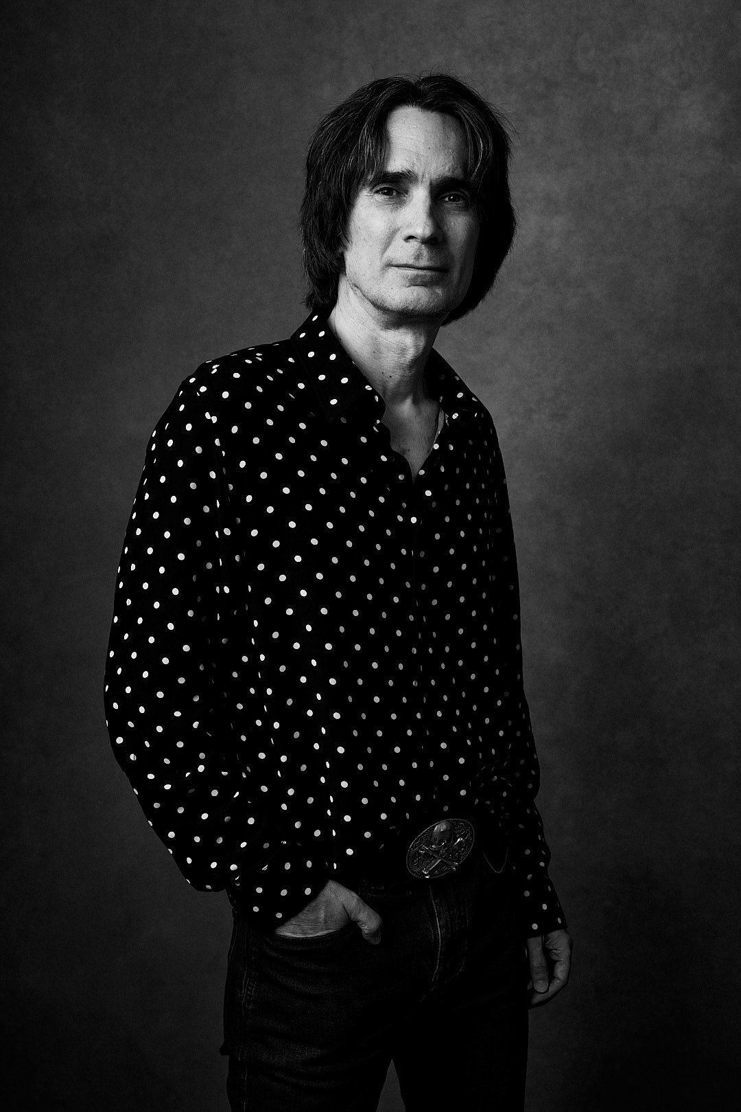 Black and white portrait of a man with shoulder-length hair wearing a polka dot shirt, standing against a plain dark background, with a relaxed pose and one hand in his pocket.
