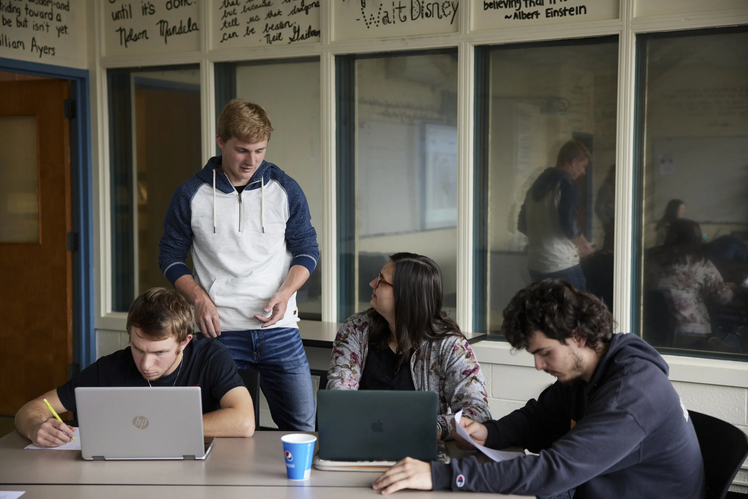 Group of four young adults working and studying together in a classroom; two are seated at a desk with laptops, one is standing and talking, and one is sitting away from the group.