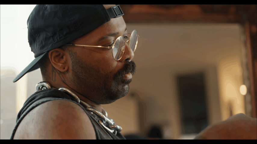 A person wearing a backwards black cap, round glasses, a chain necklace, and sleeveless shirt looking at something in their hands.