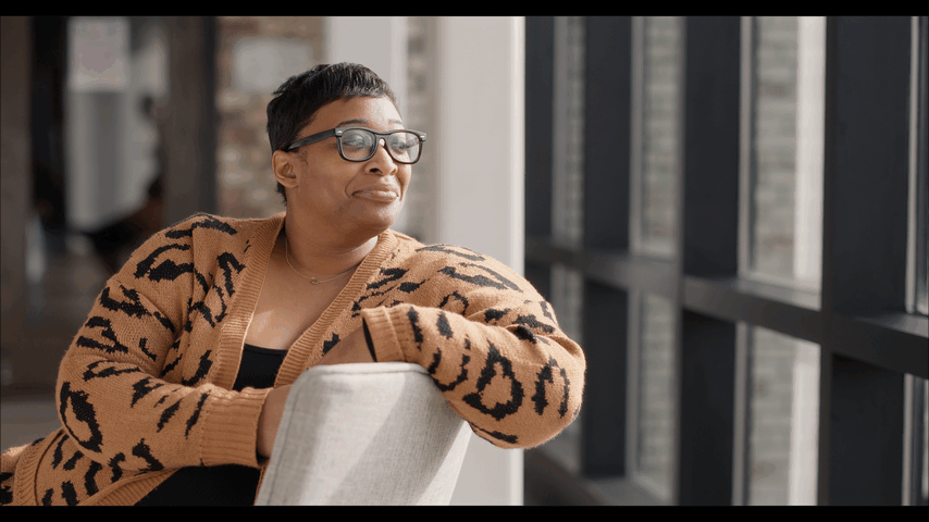Woman with short hair and glasses sitting by window, looking outside and enjoying a moment.