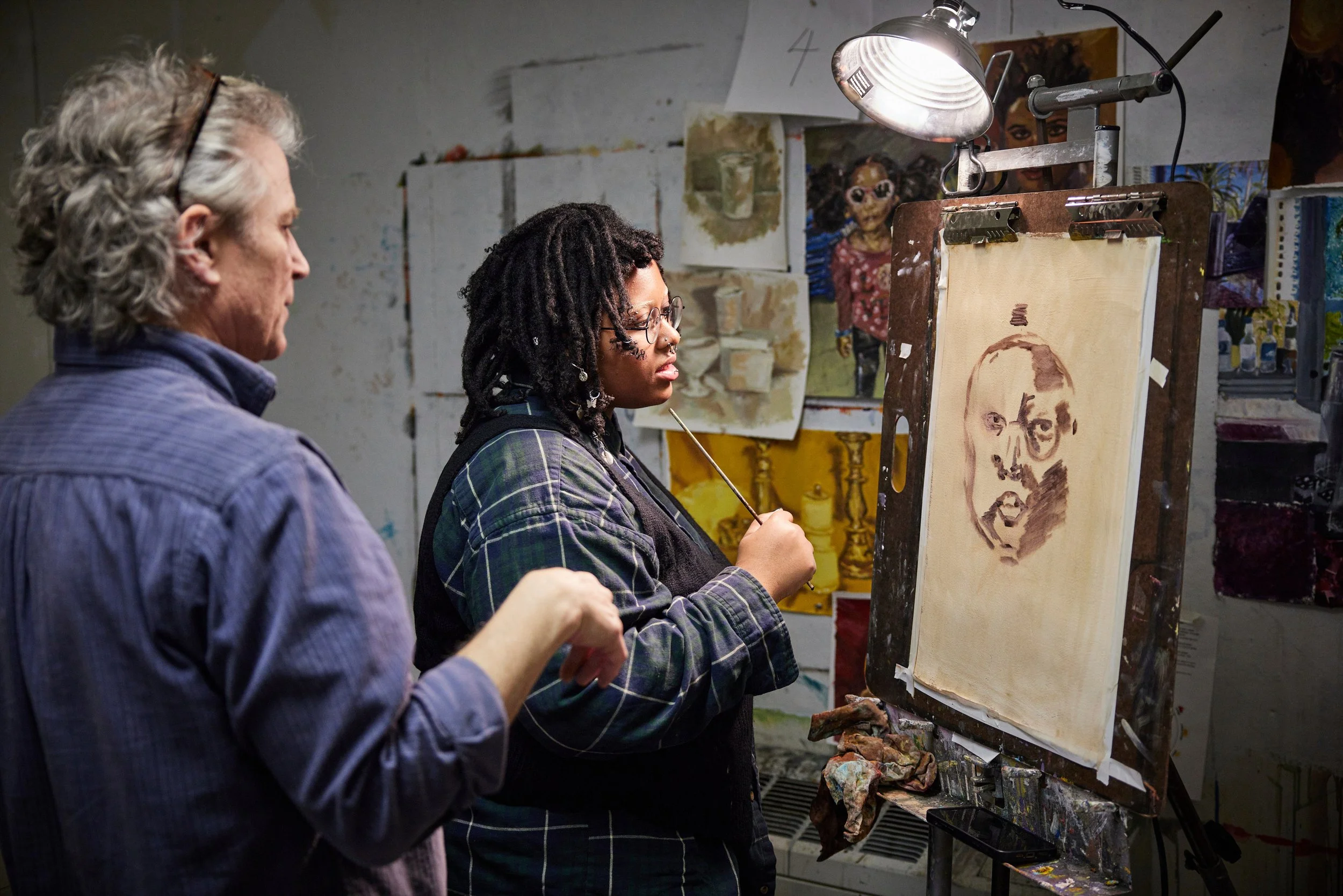 An artist and a man observing a portrait painting in progress. The artist is holding a brush and is working on a sketch of a woman's face on a canvas, with various paintings and sketches in the background.
