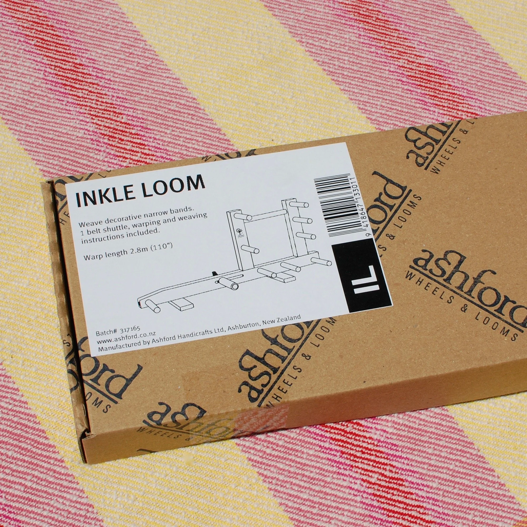 Ashford Inkle loom in its box. It rests on a handwoven pink and yellow striped textile