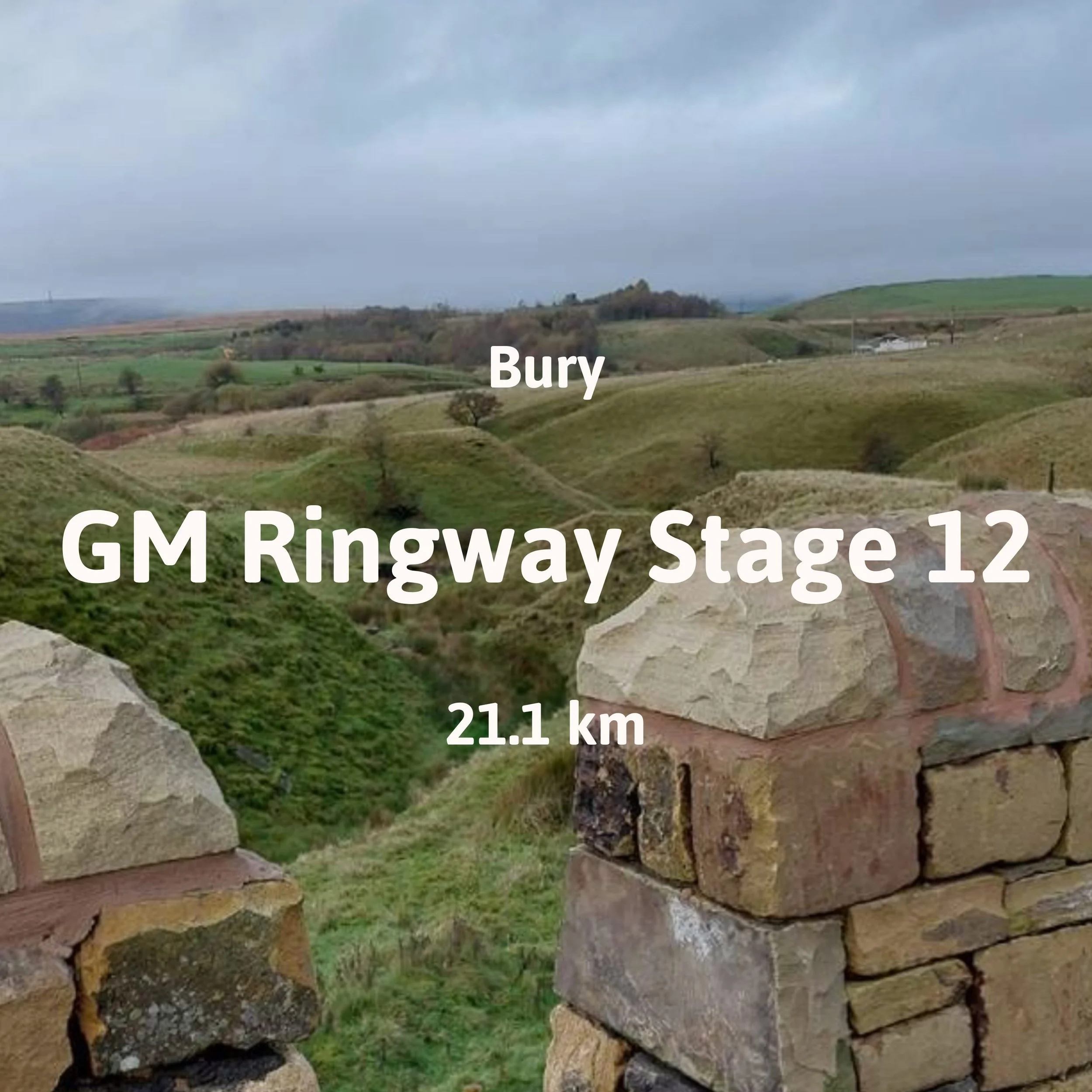 Rolling green hills and small intersecting valleys. A gap through a stone wall is in the foreground along the GM Ringway.
