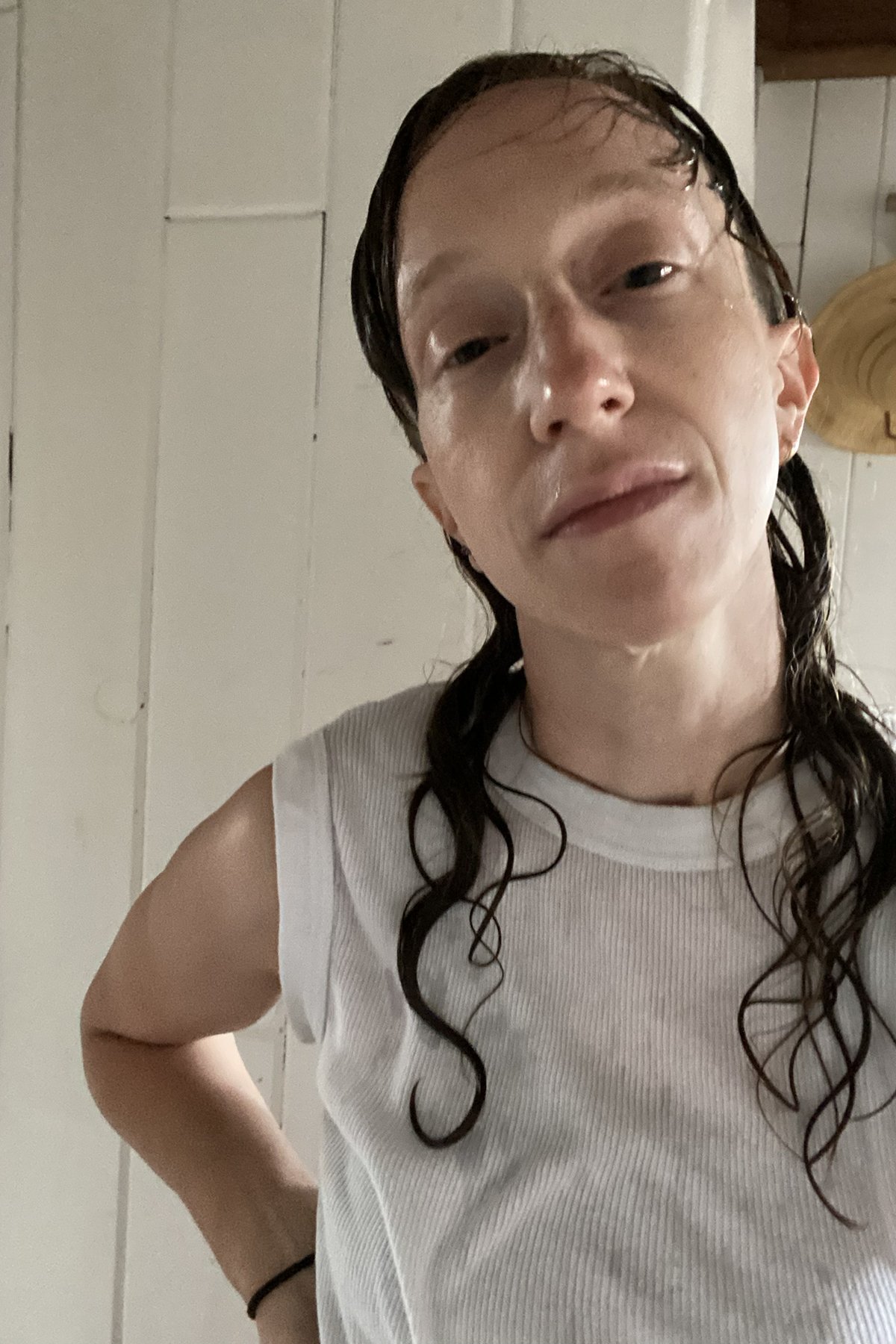 Un portrait photo montrant l'artiste, Amalle Dublon, dans une camisole de sport blanche, la peau blanche et moite, de longues ondulations plaquées entourant les côtés rasés de près. 