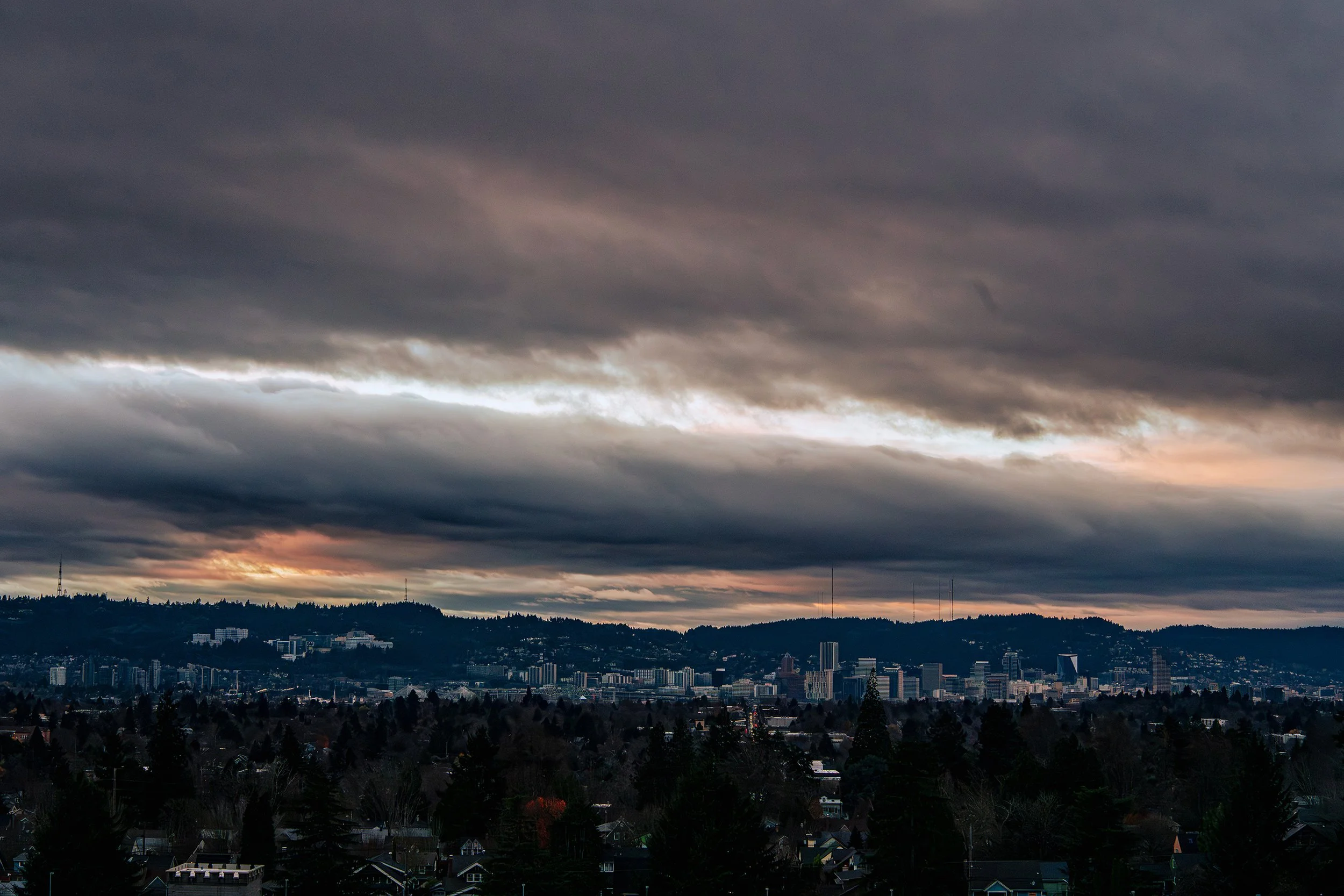 Overcast Portland cityscape with dark storm clouds and a setting sun over a downtown skyline, with residential neighborhoods in the foreground.