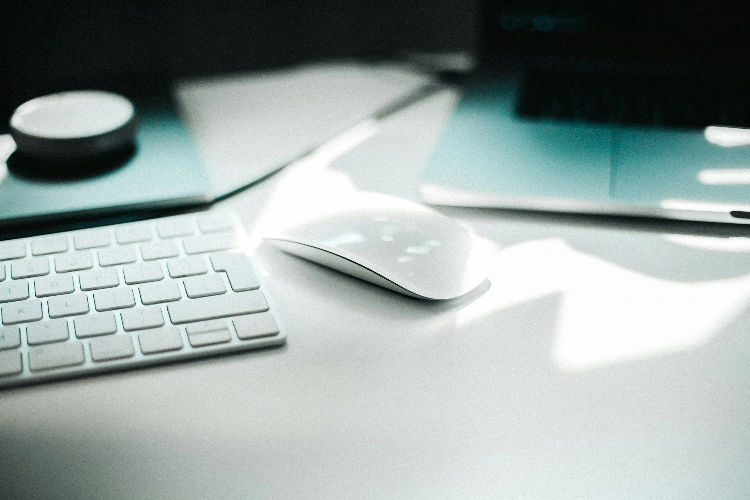 Close-up of a white computer keyboard and a white wireless mouse on a white desk with a computer monitor in the background.