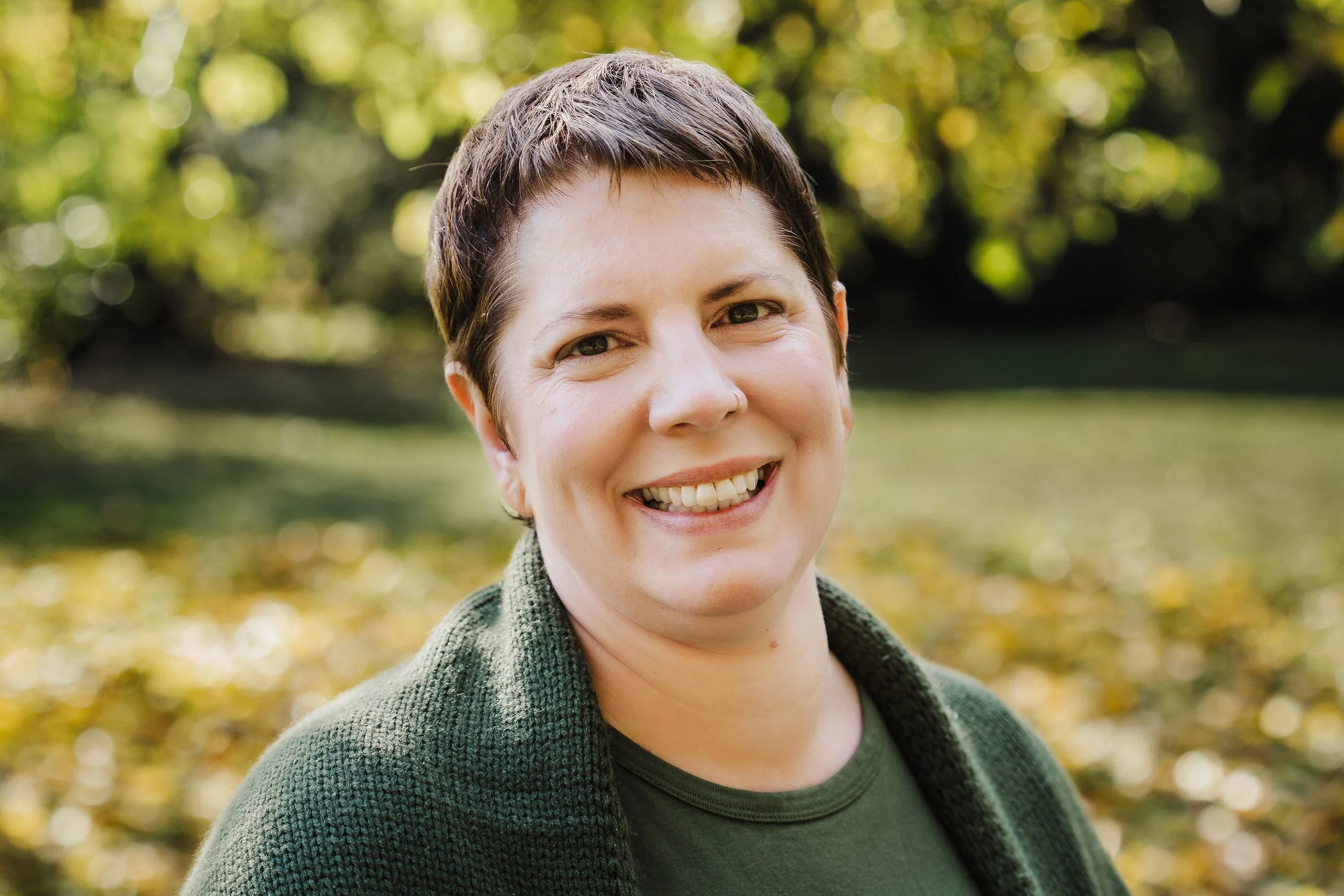 Image of a smiling doula in Portland with short brown hair and dimples standing outside in a green sweater.