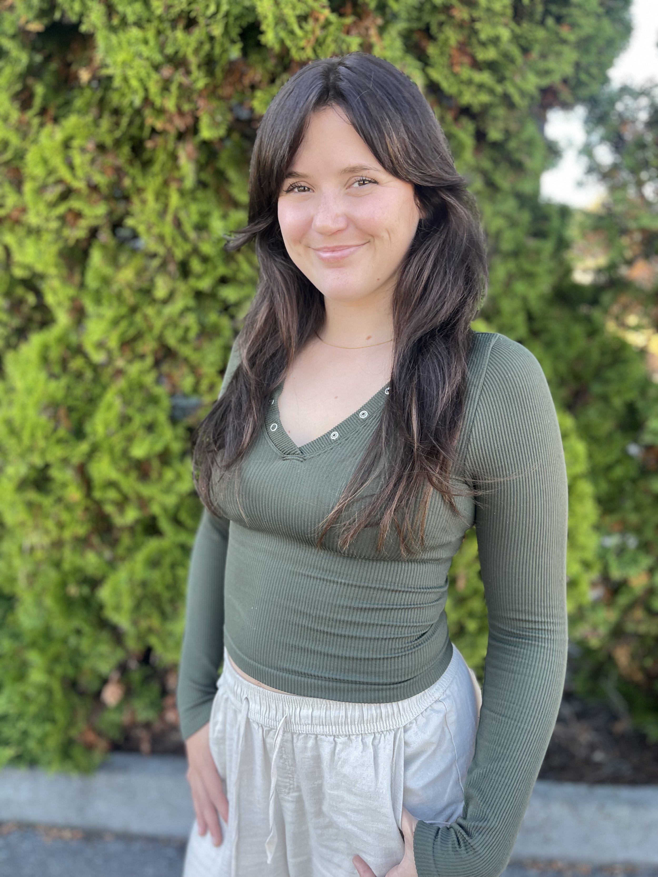 Image of a smiling dark haired doula in a green henley outside in Whidbey Island, WA